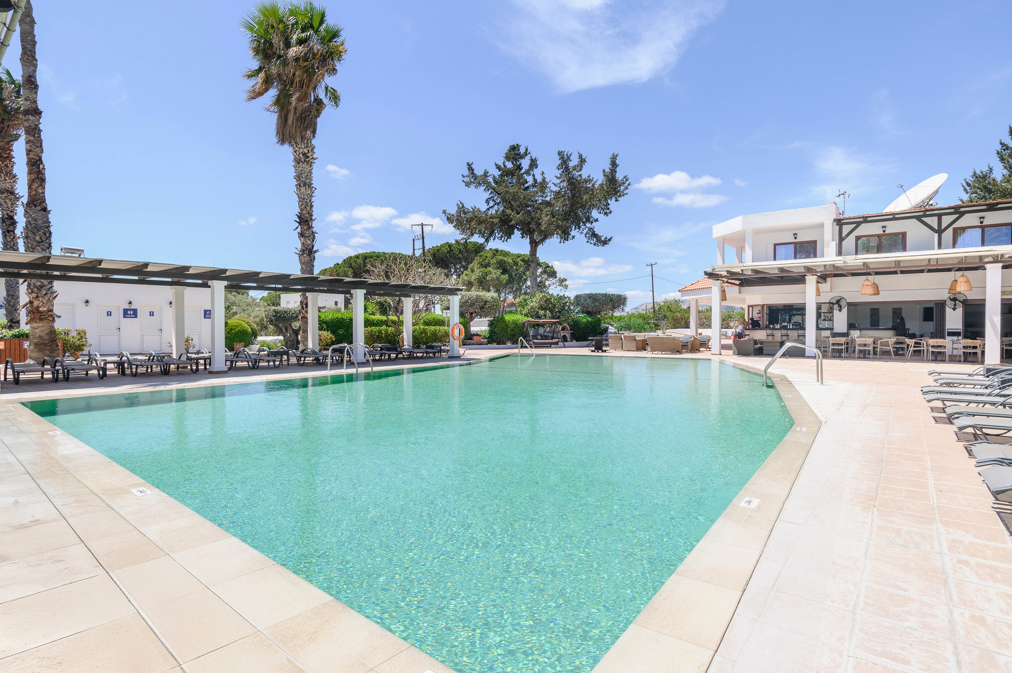 A large outdoor swimming pool is the central focus of this resort scene. The pool is surrounded by tiled decking, sun loungers, and shaded pergolas. Palm trees and trimmed greenery add a tropical touch under a bright blue sky with scattered clouds. A white two-story building with balconies and an open-air dining area frames the background, suggesting a relaxing vacation setting.