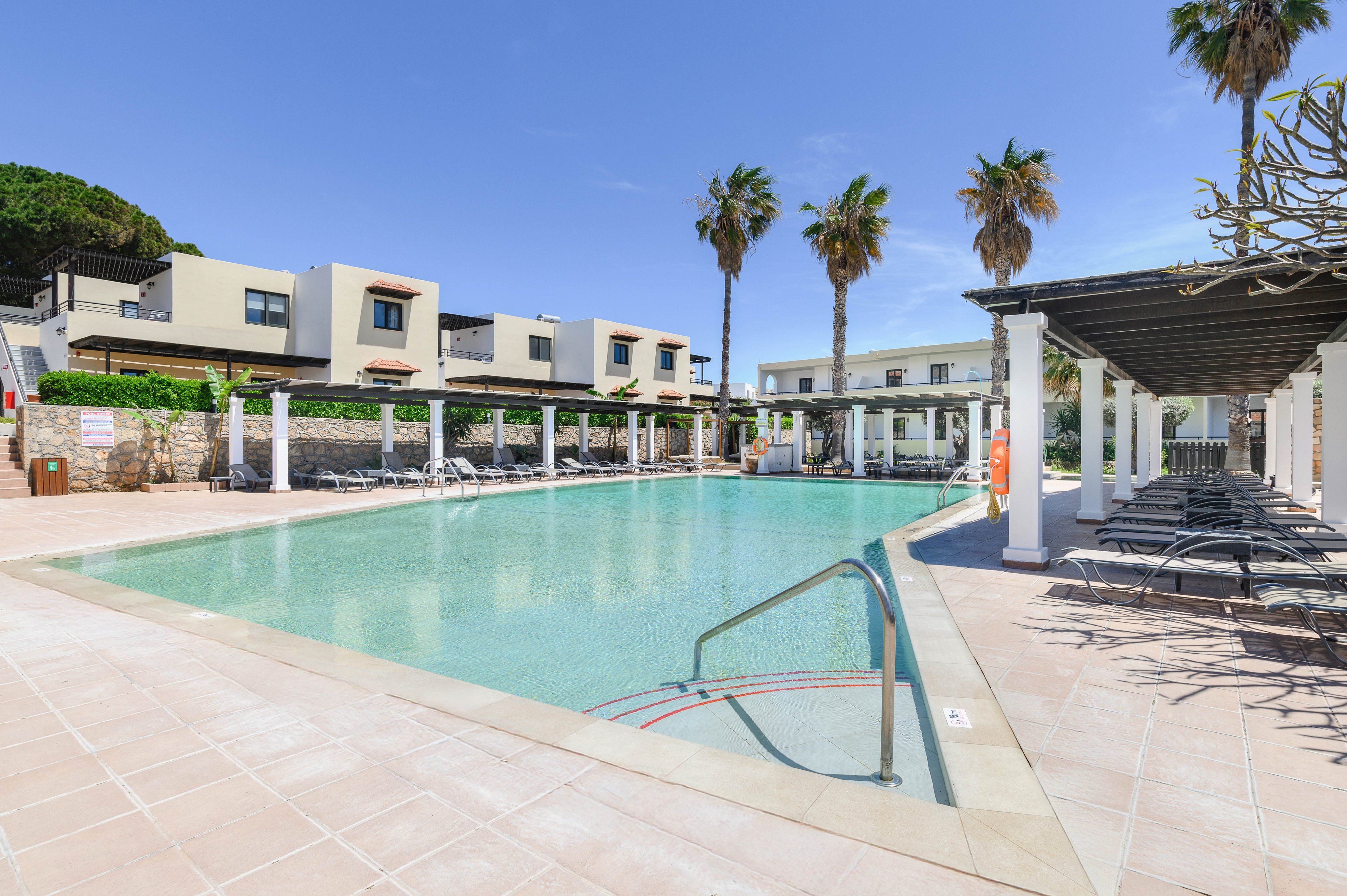 A large, clear swimming pool sits at the center of a modern resort complex. Contemporary low-rise buildings with flat roofs line the background, framed by tall palm trees. Sun loungers and shaded pergolas run along the poolside, suggesting a relaxing vacation setting. The bright daylight and clean architectural lines create a calm, minimalist atmosphere.