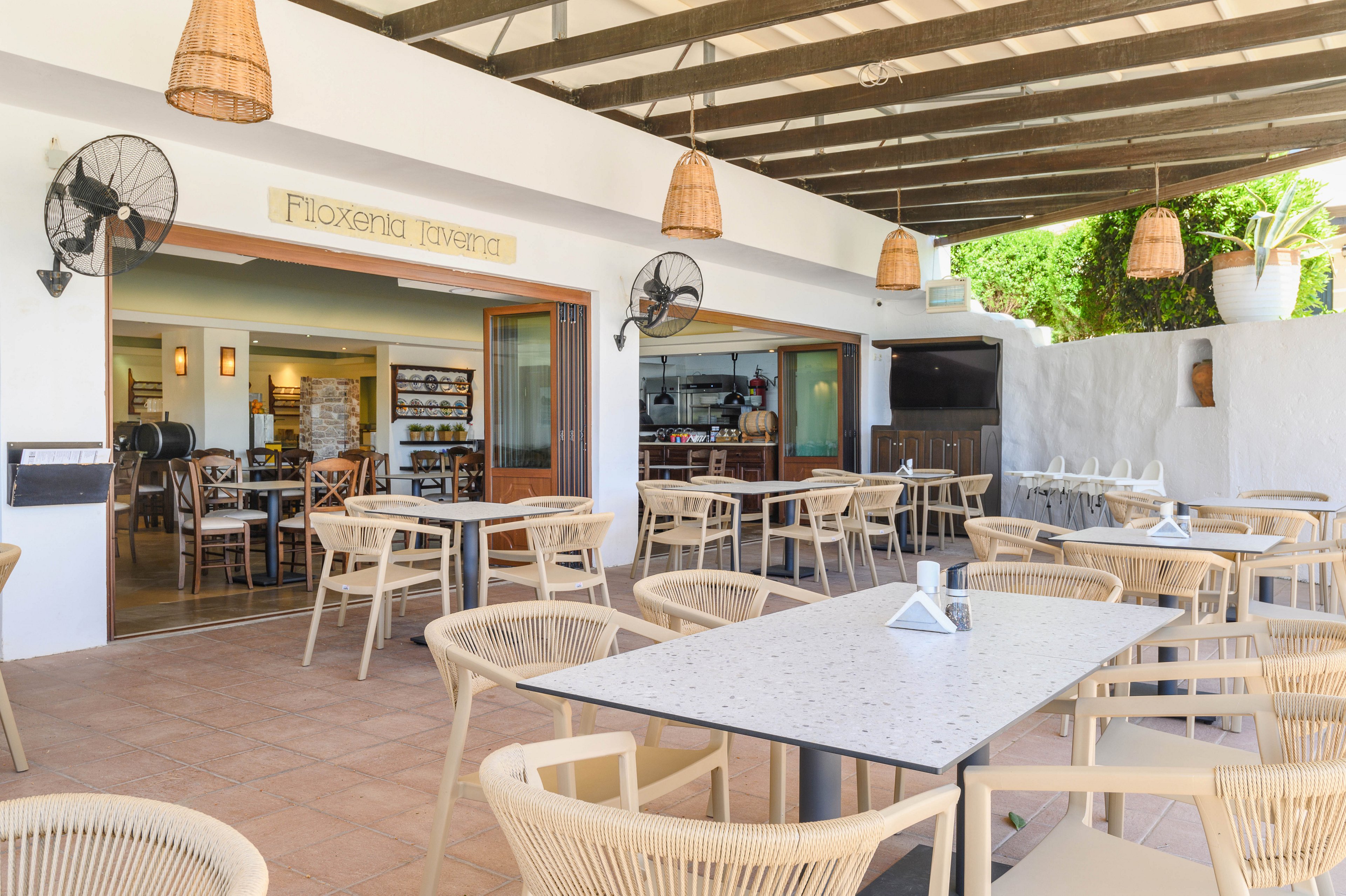 An open-air cafe with light wicker chairs and tables sits under a wooden pergola. The interior of the restaurant is visible through large openings, showing shelves, a bar area, and warm lighting. Ceiling fans and woven pendant lamps hang above the terrace, while greenery and whitewashed walls enhance the Mediterranean feel. The space appears calm and ready for guests, with neatly arranged furniture and clean surfaces.