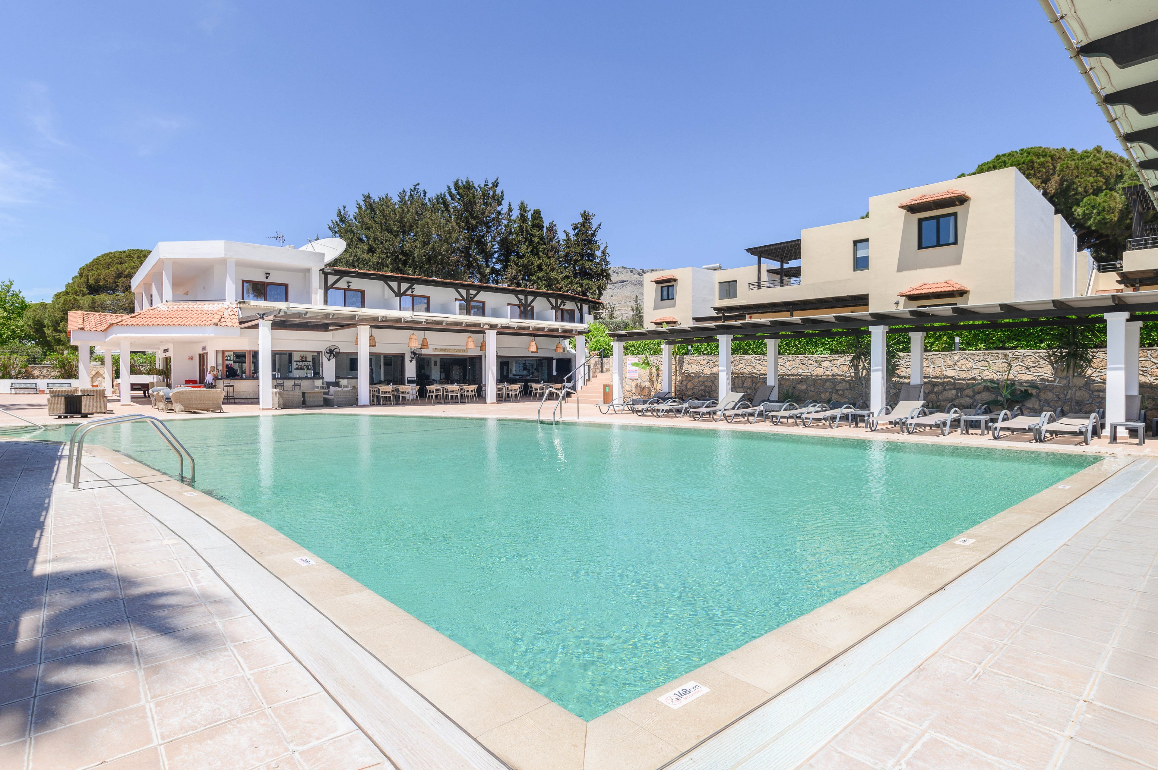 A large turquoise swimming pool dominates the foreground of a modern resort complex. White and beige buildings with flat roofs and balconies surround the pool area under a clear blue sky. Sun loungers line the poolside beneath a shaded pergola, suggesting a relaxing vacation setting. The bright daylight and clean architectural lines create a calm, inviting atmosphere.