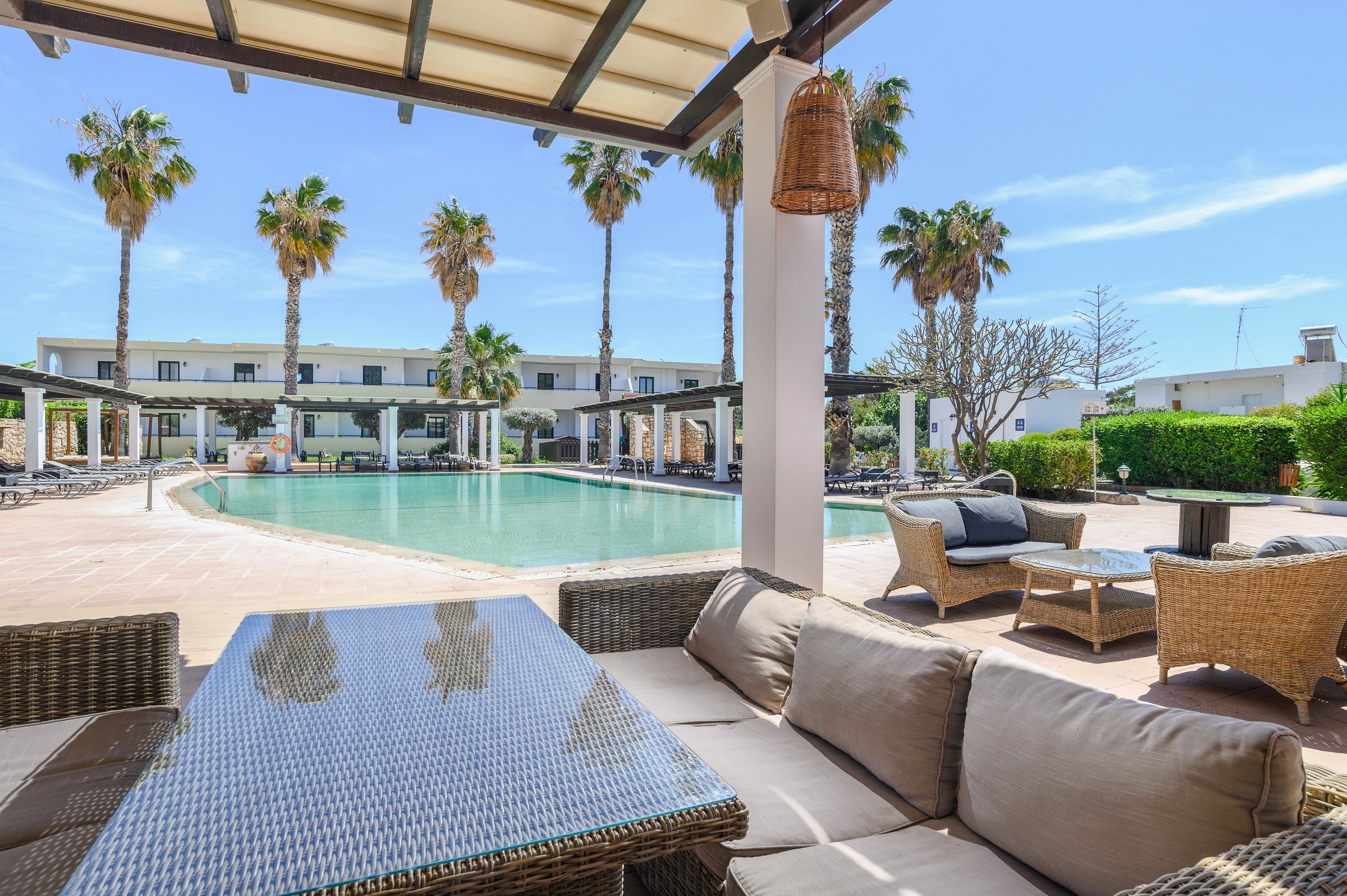 An inviting resort pool area features a shaded lounge with wicker sofas and a glass-topped table in the foreground. Beyond the covered terrace, a large swimming pool stretches out, surrounded by sun loungers and umbrellas. A modern low-rise hotel building frames the background, with tall palm trees adding a tropical atmosphere. The bright blue sky and sunlight create a relaxed, vacation-like setting ideal for outdoor relaxation.