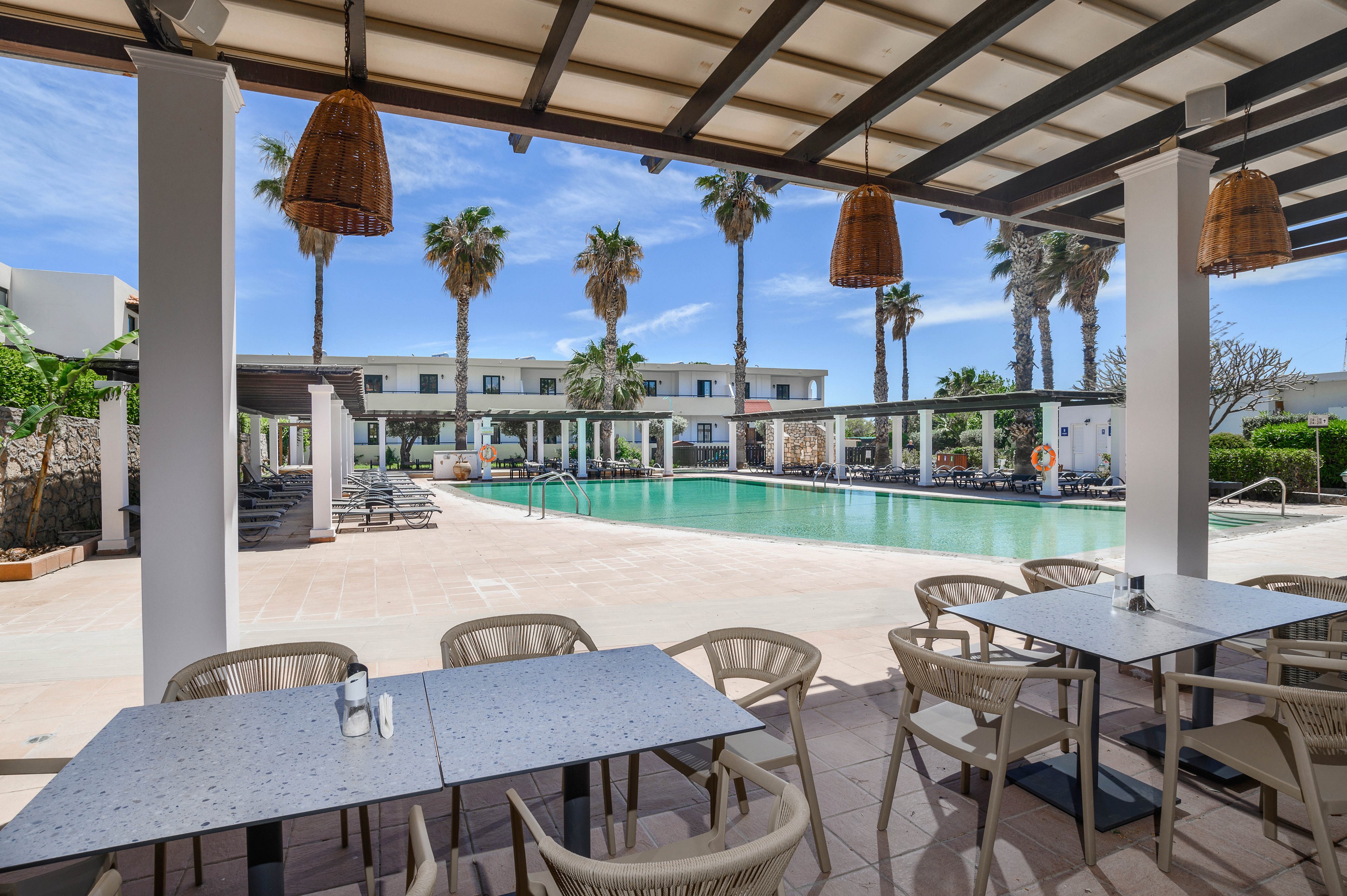 An open-air dining area overlooks a large resort swimming pool framed by tall palm trees. White tables and plastic chairs sit under a shaded pergola with hanging wicker lamps. In the background, sun loungers and umbrellas line the pool deck near a modern low-rise building. The scene is bright and sunny, suggesting a warm vacation destination with clear blue skies.