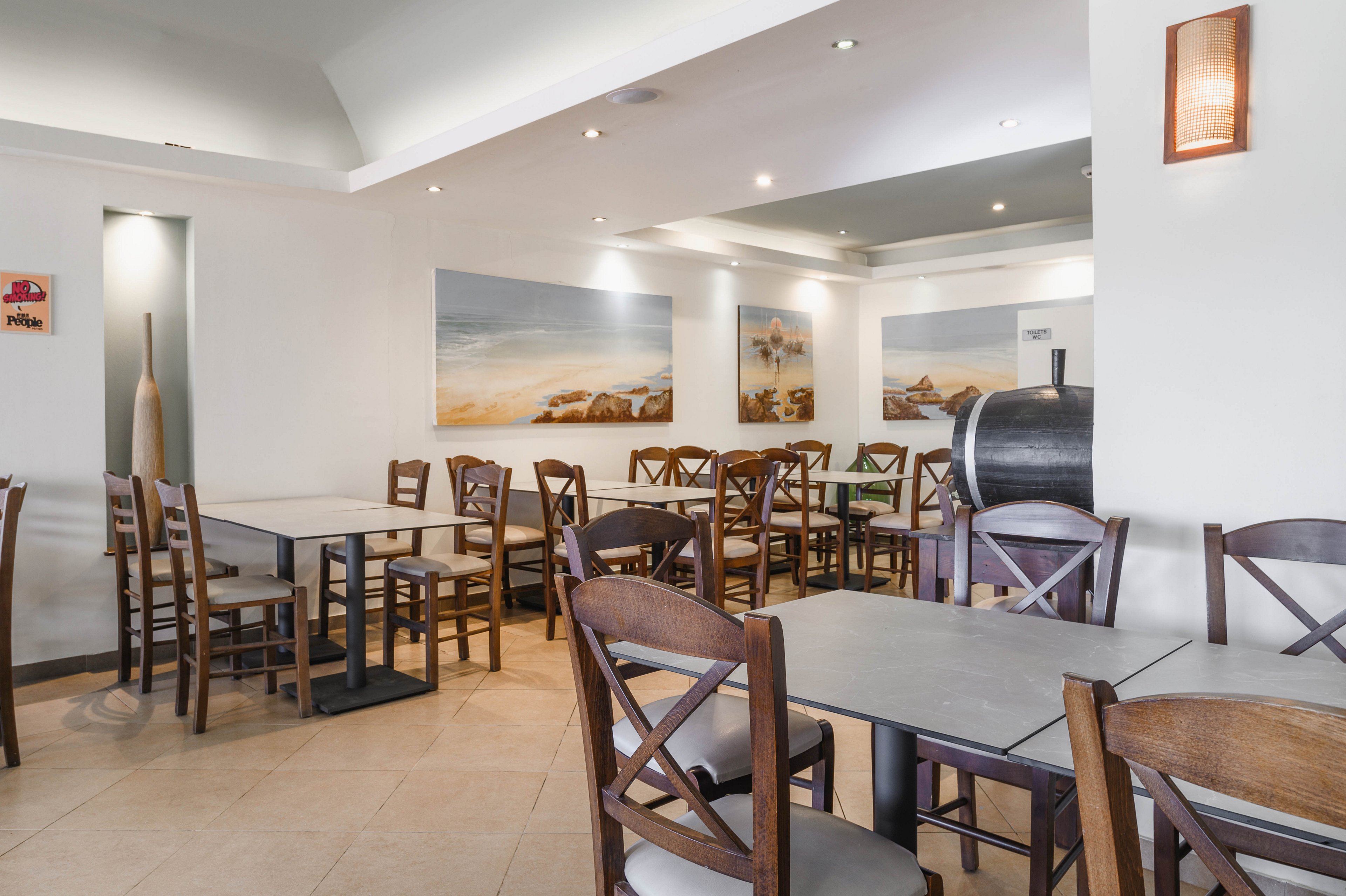A clean, modern restaurant dining room is shown with neatly arranged wooden chairs and gray tabletops. The space is empty of people, emphasizing the orderly layout and ample seating. Soft ceiling lights and wall sconces create a warm, inviting atmosphere. Minimalist wall art with coastal scenes and neutral tones enhances the contemporary design.