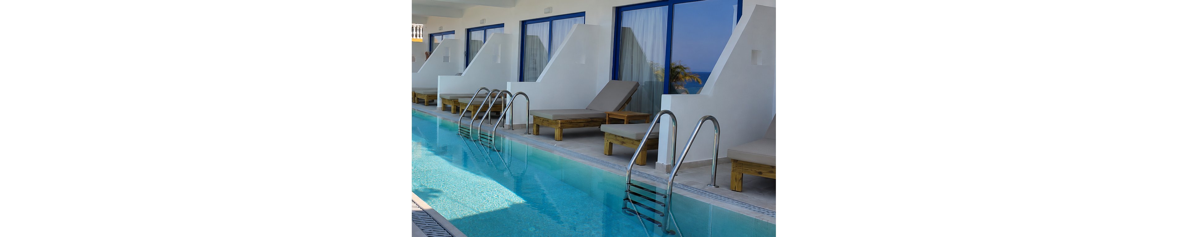 A row of contemporary hotel suites opens directly onto a narrow swimming pool. Wooden sun loungers with beige cushions line the poolside beneath white architectural walls. Blue-framed glass doors and windows reflect the bright daylight and hint at an ocean view beyond. Stainless steel pool ladders lead into clear turquoise water, creating a relaxing vacation atmosphere.