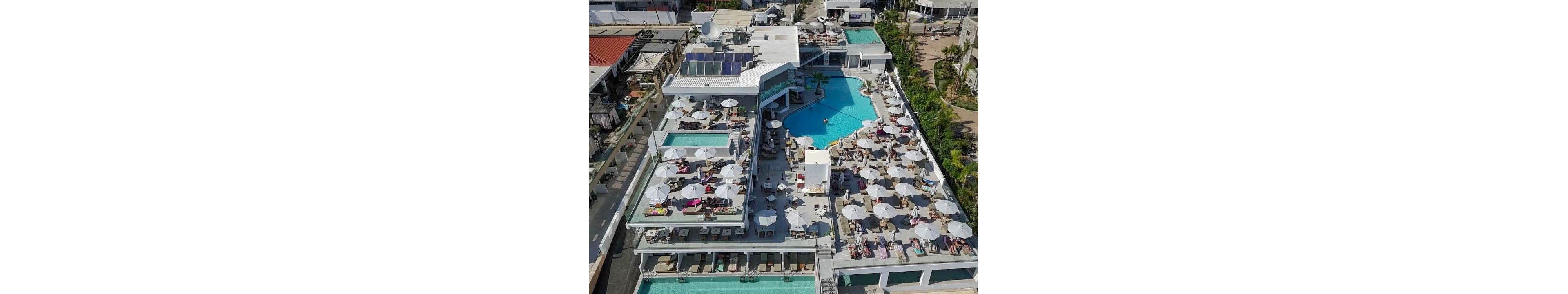 A large multi-level resort complex with several outdoor swimming pools is captured from an aerial perspective. Numerous lounge chairs and white umbrellas surround the pools, with many guests sunbathing and relaxing. The architecture features modern white buildings, terraces, and glass railings. Palm trees and landscaped walkways frame the property, suggesting a warm coastal or tropical vacation destination.