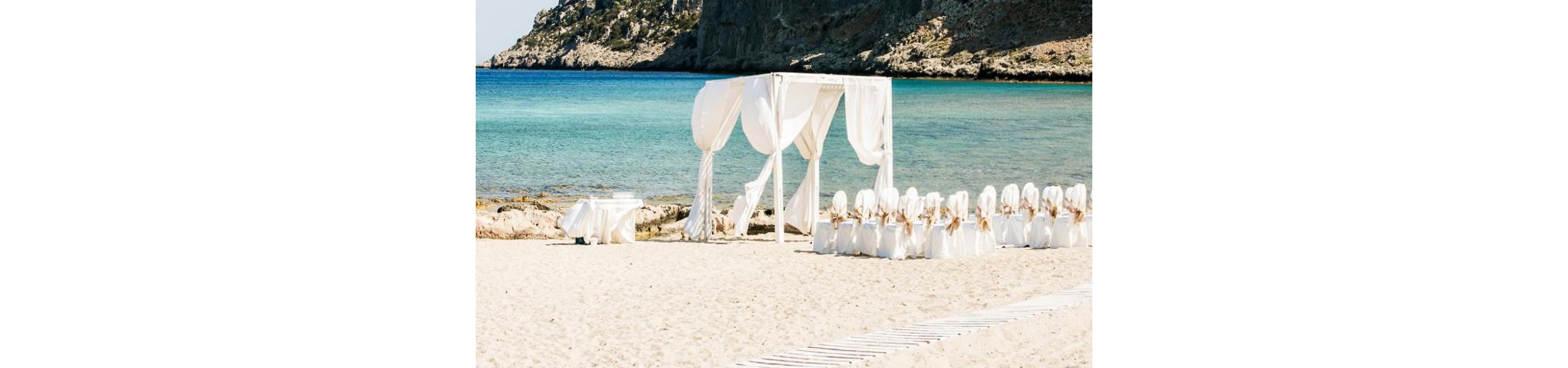 A white-draped wedding arch stands on a sandy beach beside clear turquoise water. Rows of chairs covered in white fabric are arranged neatly for guests, facing the ceremony area. Rocky cliffs and a calm sea create a scenic coastal backdrop. The overall scene feels bright, romantic, and serene under clear daylight.