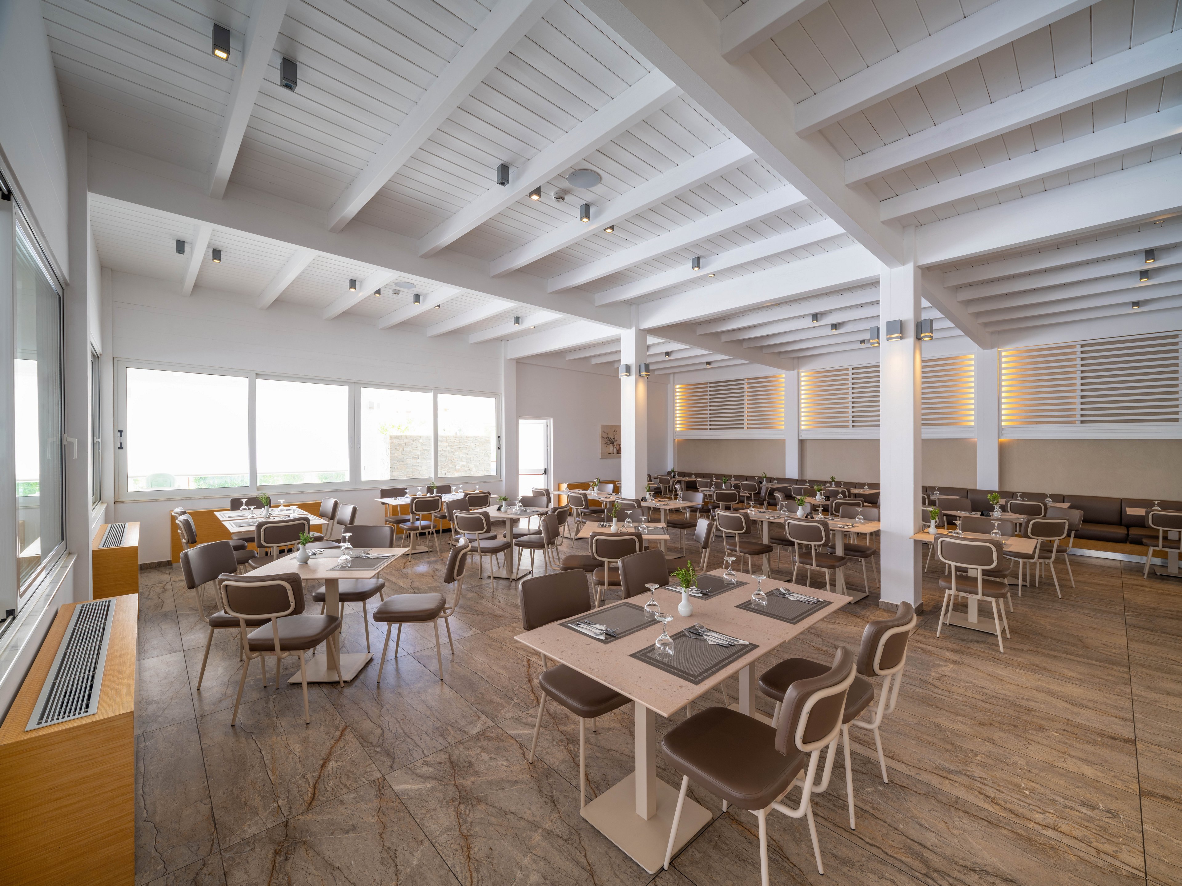 Spacious contemporary restaurant interior with neatly arranged tables and chairs. Large windows fill the room with natural light, highlighting the light wood furniture and clean lines. The ceiling features exposed white beams and recessed lighting, creating an airy atmosphere. Neutral tones and minimalist decor give the dining area a calm, modern feel.
