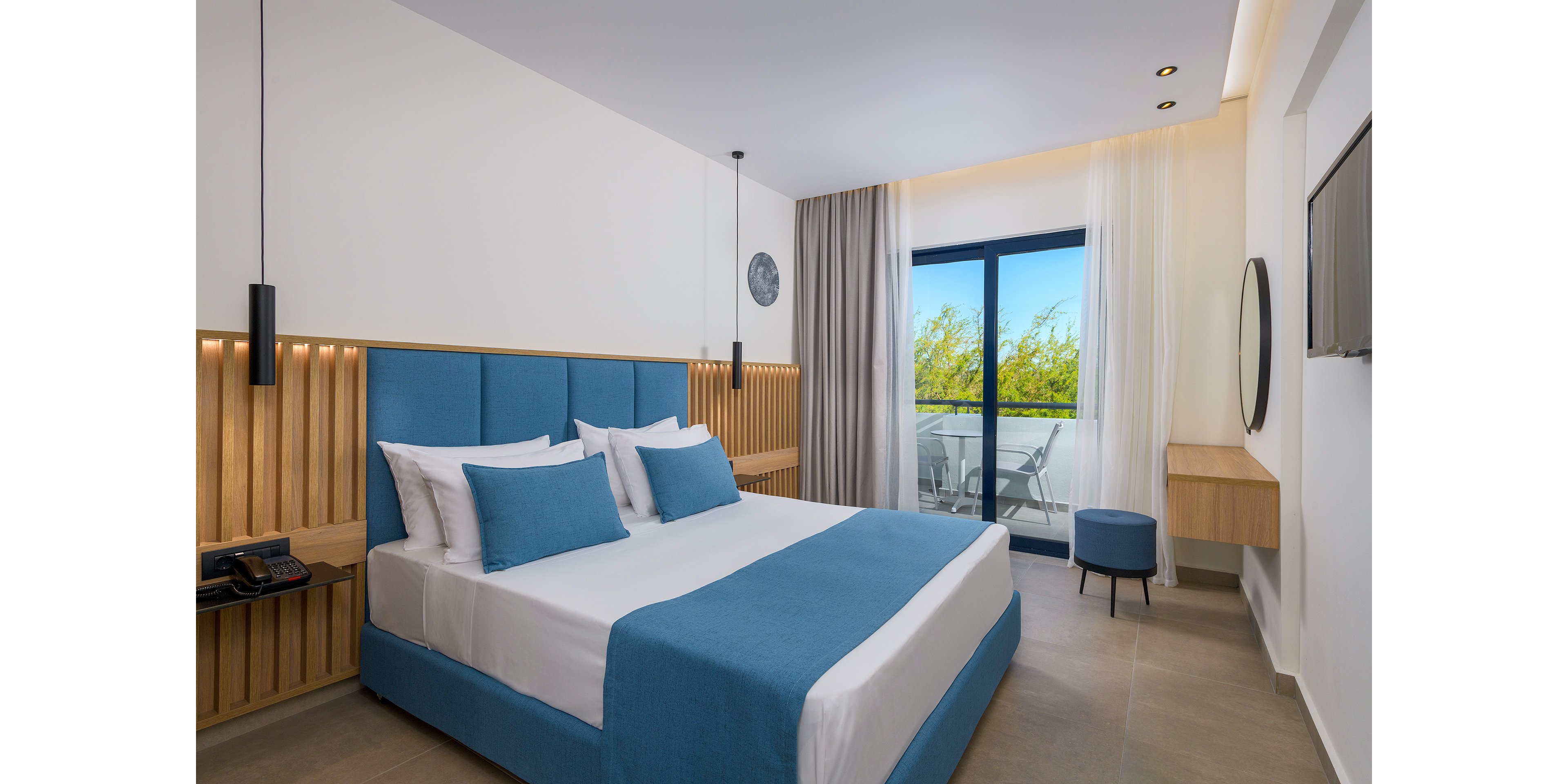 A contemporary hotel room featuring a neatly made bed with blue and white bedding. The room includes wooden paneling, pendant lights, and a balcony with a view of greenery. The decor is minimalistic with a soothing color palette of blue and natural wood tones.
