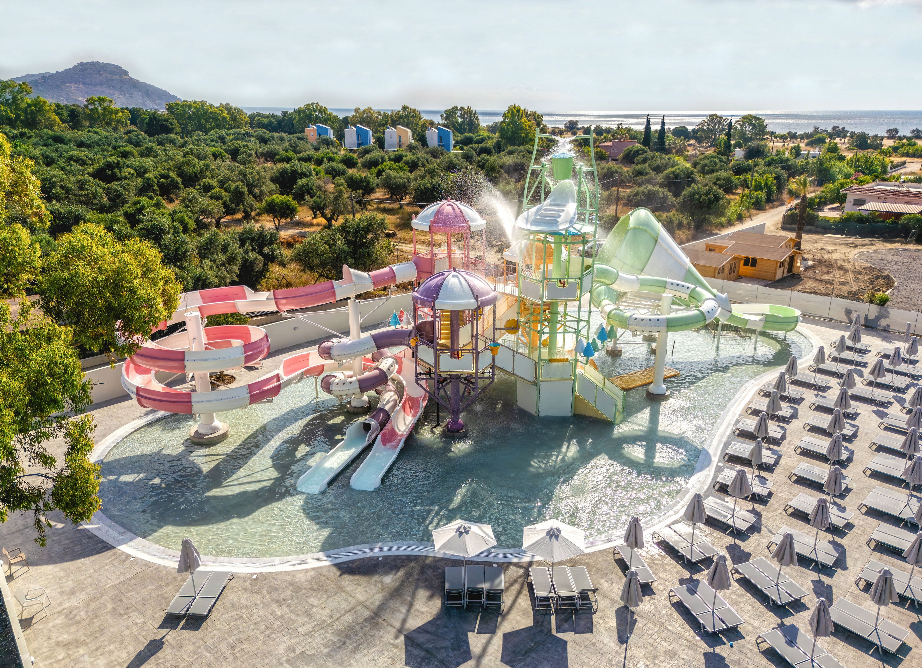 A vibrant water play area with multiple colorful slides and splash structures is shown at a resort. The shallow pool surrounds towers, tunnels, and curved slides in pink, red, green, and white tones. Rows of sun loungers with umbrellas line the deck around the water park, suggesting a family-friendly leisure space. In the background, lush greenery, scattered buildings, and a distant coastline create a scenic holiday setting.