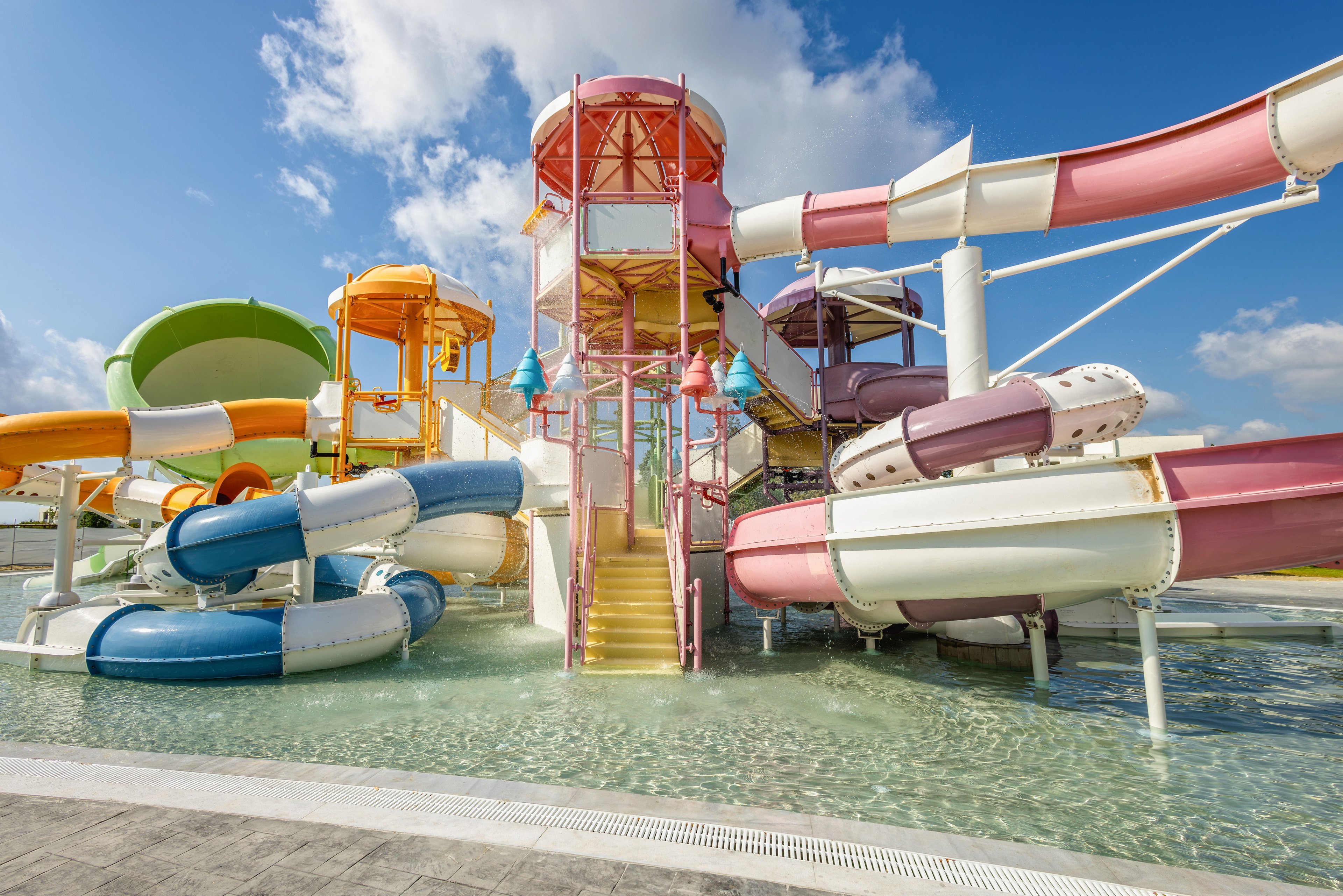A vibrant water park features a central play tower surrounded by multiple twisting slides in bright colors. The structure rises from a shallow pool of clear water under a sunny blue sky with scattered clouds. Yellow stairs lead up through the tower, while curved tubes in orange, pink, green, and blue create a dynamic, playful scene. The overall setting suggests a fun, family-friendly outdoor attraction designed for warm weather recreation.