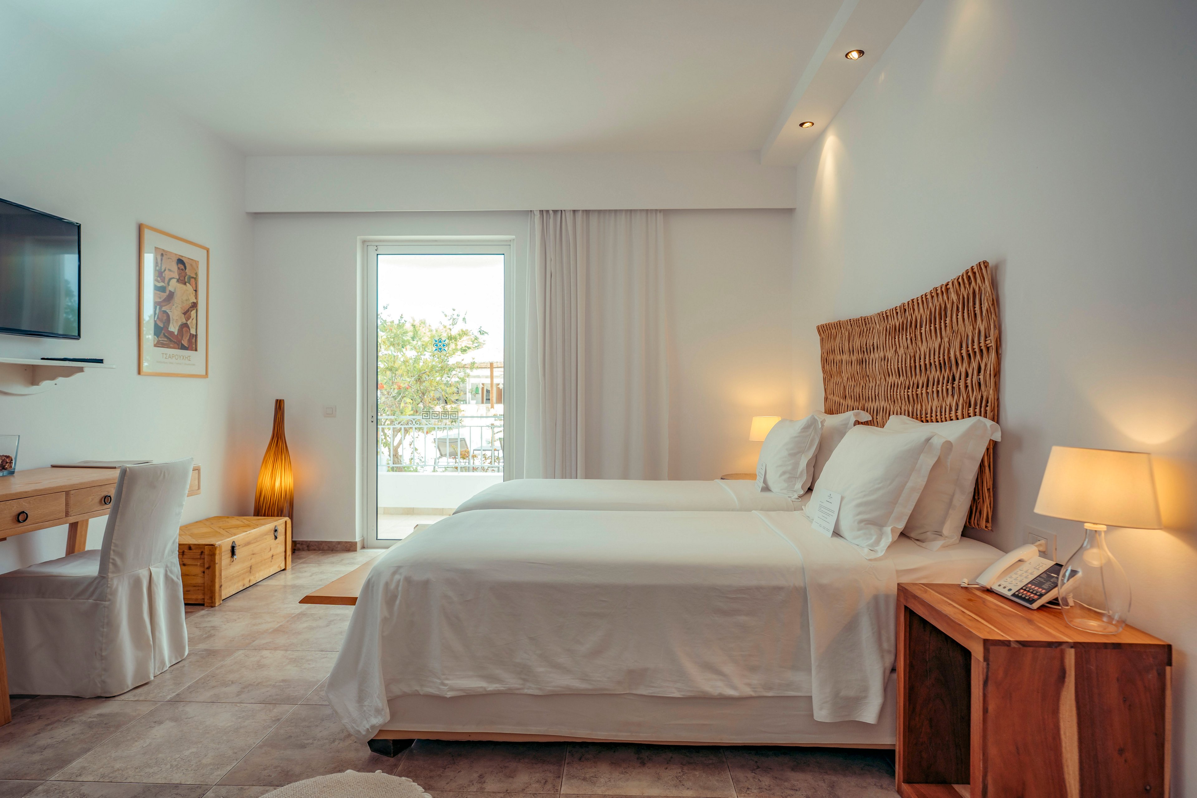 A modern hotel-style bedroom features a neatly made double bed with white linens and a woven headboard. The room is decorated in warm neutral tones with wooden furniture, including bedside tables, a desk, and a low chest. Soft lighting from table lamps and a tall floor lamp creates a cozy atmosphere. A wall-mounted television and a framed artwork add contemporary touches. Natural light enters through a glass door leading to a balcony with a partial outdoor view.