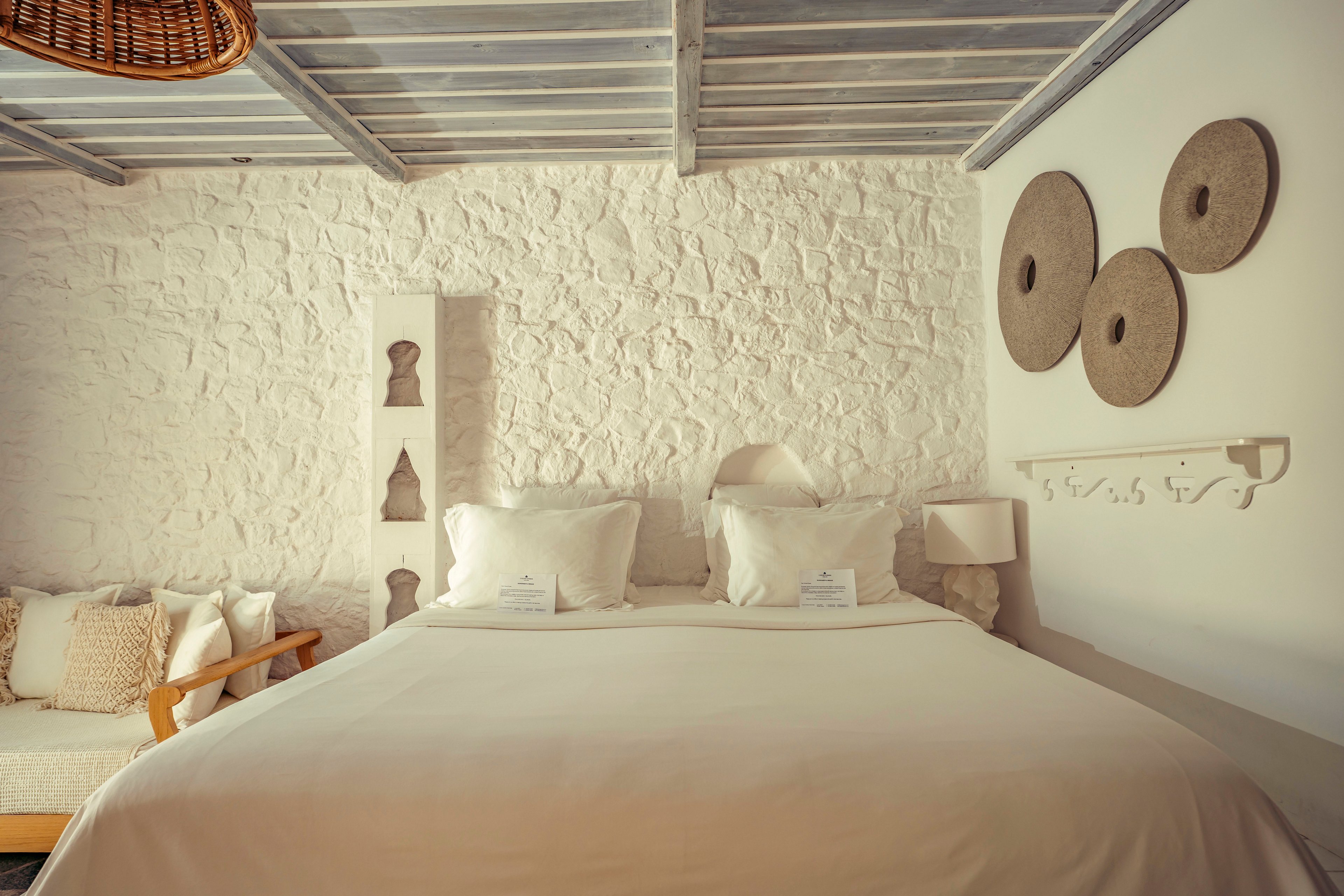 A neatly made double bed with crisp white linens is centered in a rustic bedroom. The room features textured white stone walls, a wooden ceiling with exposed beams, and soft neutral decor. A small sofa with cushions sits to the side, while a bedside lamp and decorative wall niches add character. Circular wall art and a woven light fixture contribute to the warm, minimalist aesthetic.