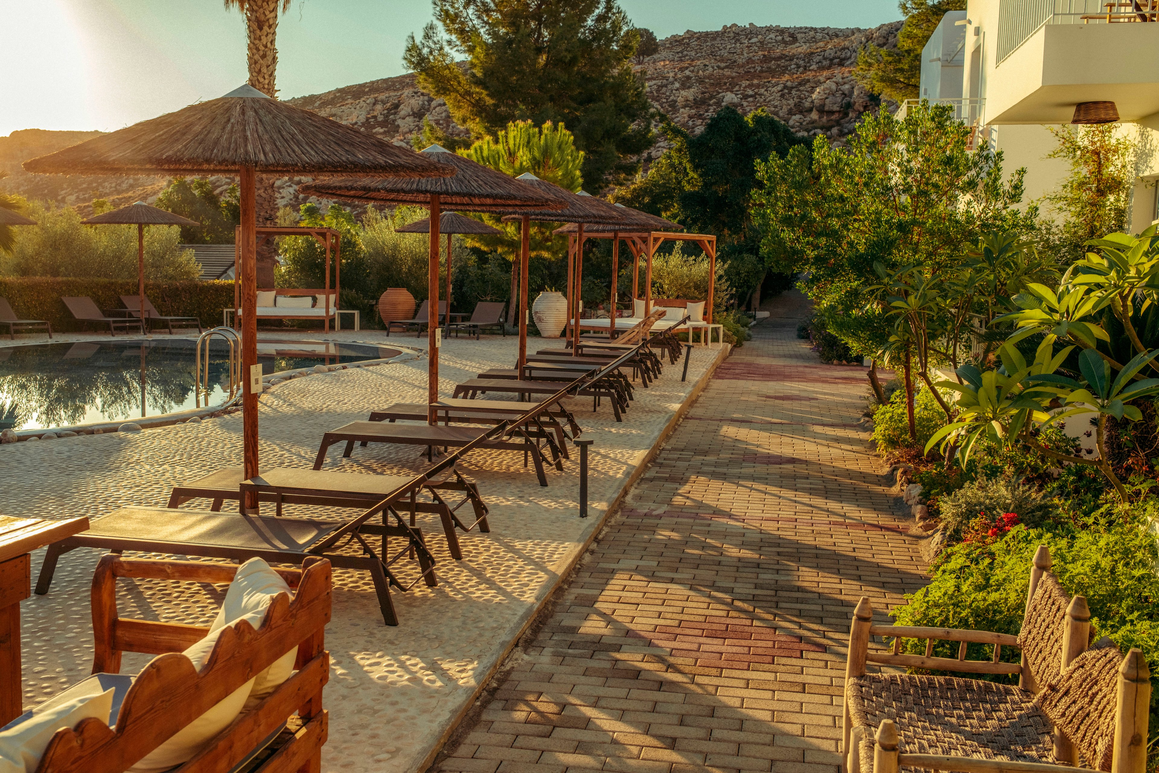 A tranquil resort pool area is shown at golden hour with rows of empty sun loungers under thatched umbrellas. The swimming pool reflects the warm sunlight, creating a calm and inviting atmosphere. A paved walkway runs alongside lush green plants and trees, leading toward the distant hillside. The scene feels peaceful and relaxed, with no people present and soft shadows stretching across the ground.