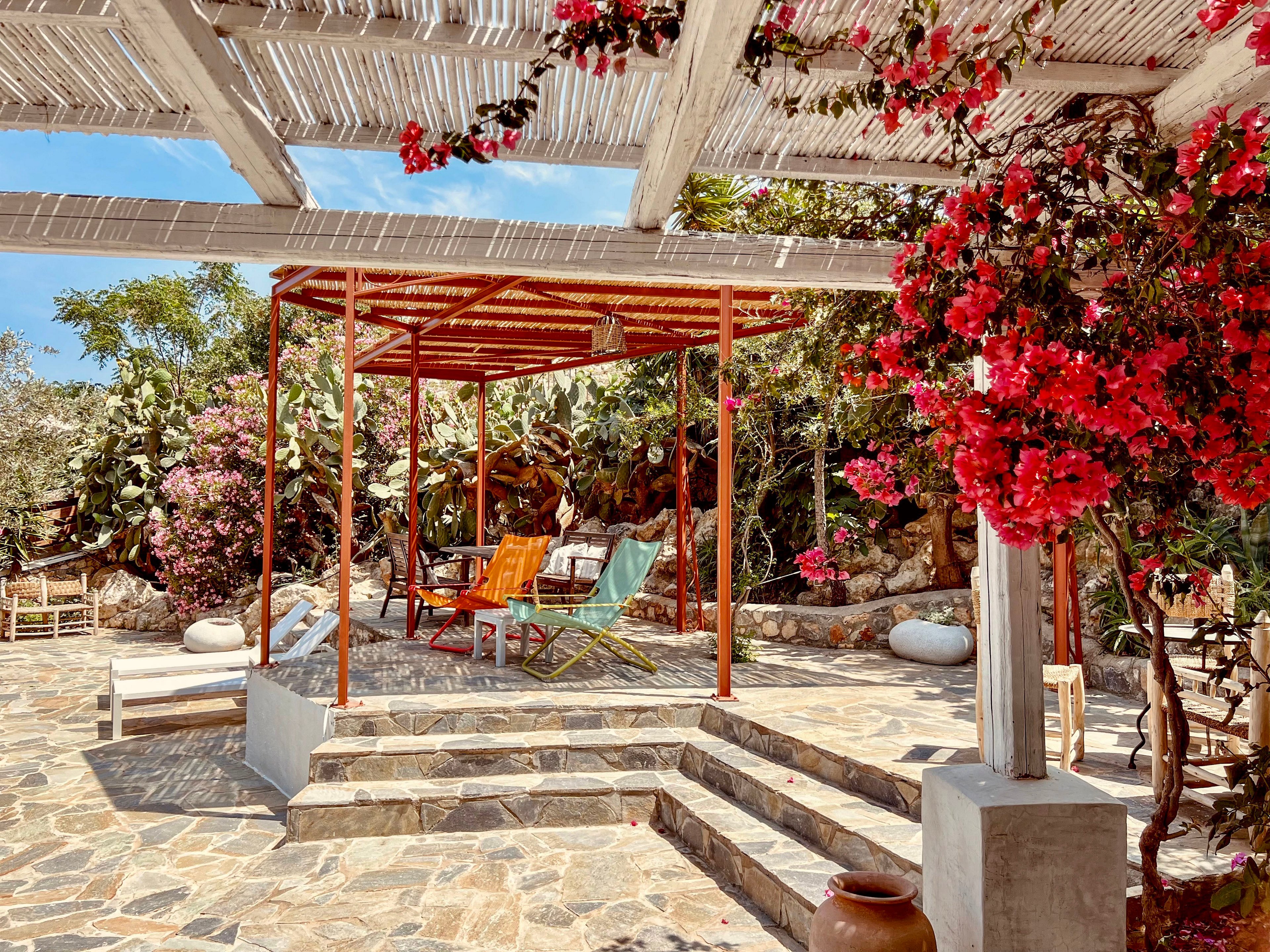 An outdoor patio features colorful lounge chairs arranged under a simple red metal pergola. The stone-paved terrace is surrounded by lush greenery, flowering bougainvillea, and rustic garden elements. Sunlight filters through a white slatted roof, casting soft shadows on the steps and seating area. The scene evokes a relaxed, summery Mediterranean atmosphere ideal for leisure and outdoor living.