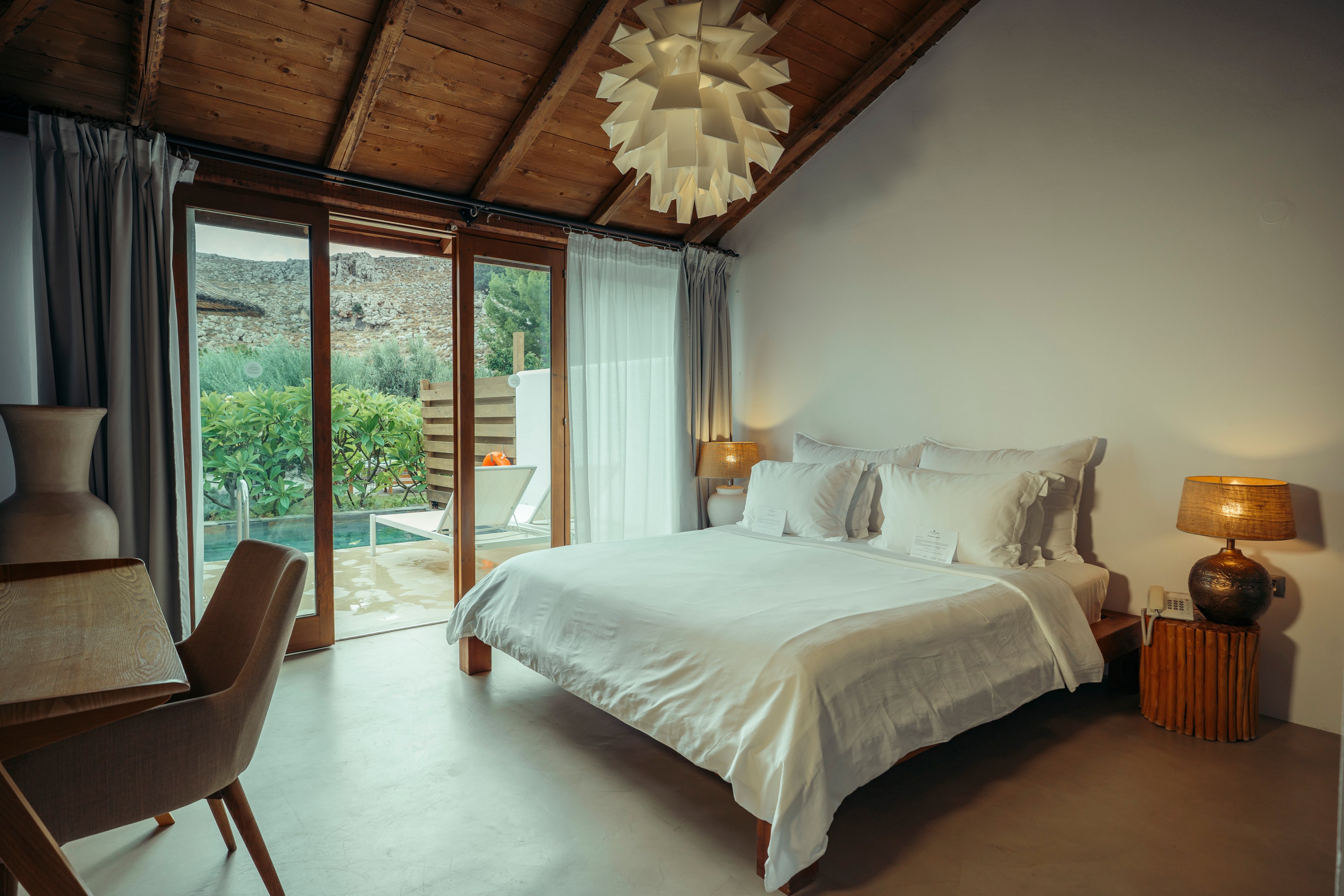 A neatly made double bed with white linens sits in a cozy resort bedroom. Large glass doors open to a private patio with greenery and a glimpse of a pool outside. Warm bedside lamps and a wooden ceiling create a soft, inviting glow. Neutral tones and minimalist furniture give the room a calm, contemporary feel.