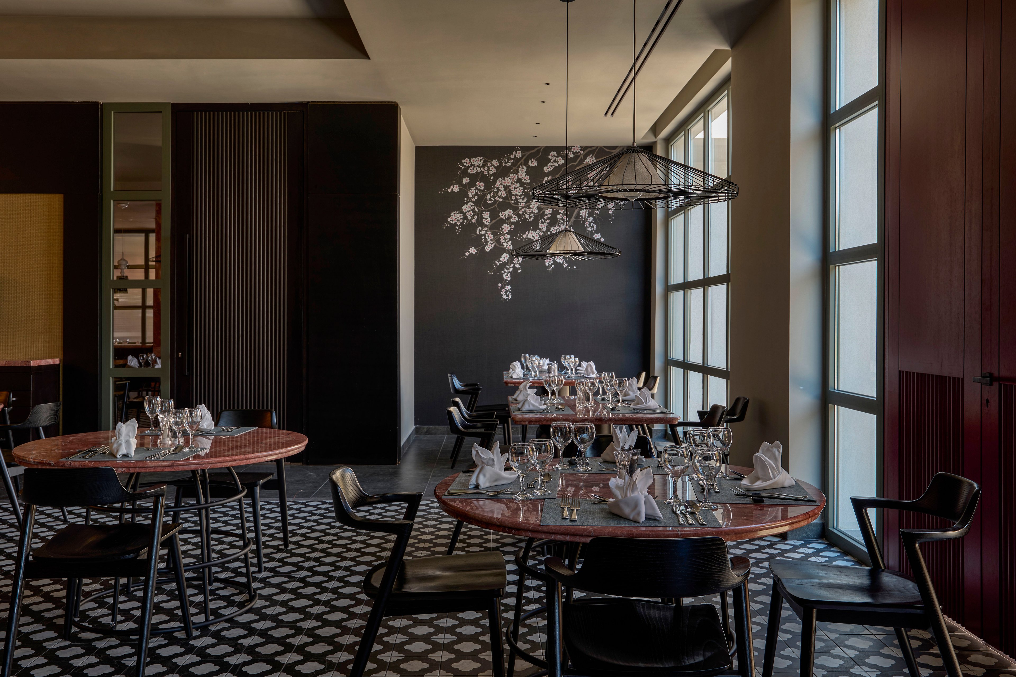 Contemporary restaurant dining room with round wooden tables set for service. Wine glasses, folded white napkins, and cutlery are neatly arranged on each table, creating a refined atmosphere. Large floor-to-ceiling windows on the right bring natural light into the dark-toned space. A decorative wall with a floral mural and geometric patterned floor add visual interest and modern style.