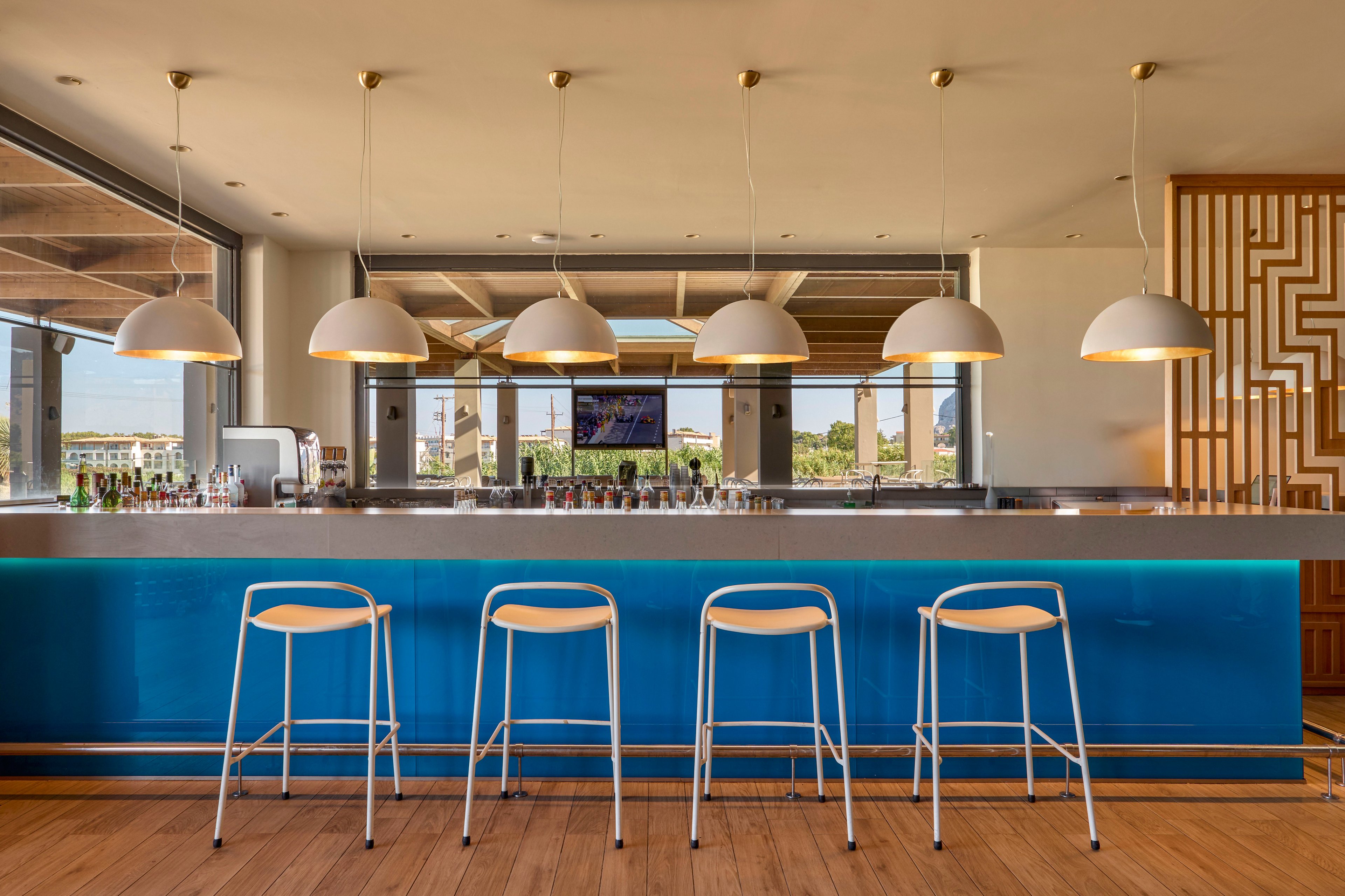 A contemporary bar interior features a long blue counter with four white bar stools neatly aligned in front. Five dome-shaped pendant lights hang above the counter, casting warm illumination on the sleek surface. Large windows behind the bar reveal an outdoor view and allow natural light to fill the space. The overall design is clean and minimal, emphasizing modern lines and a relaxed atmosphere.