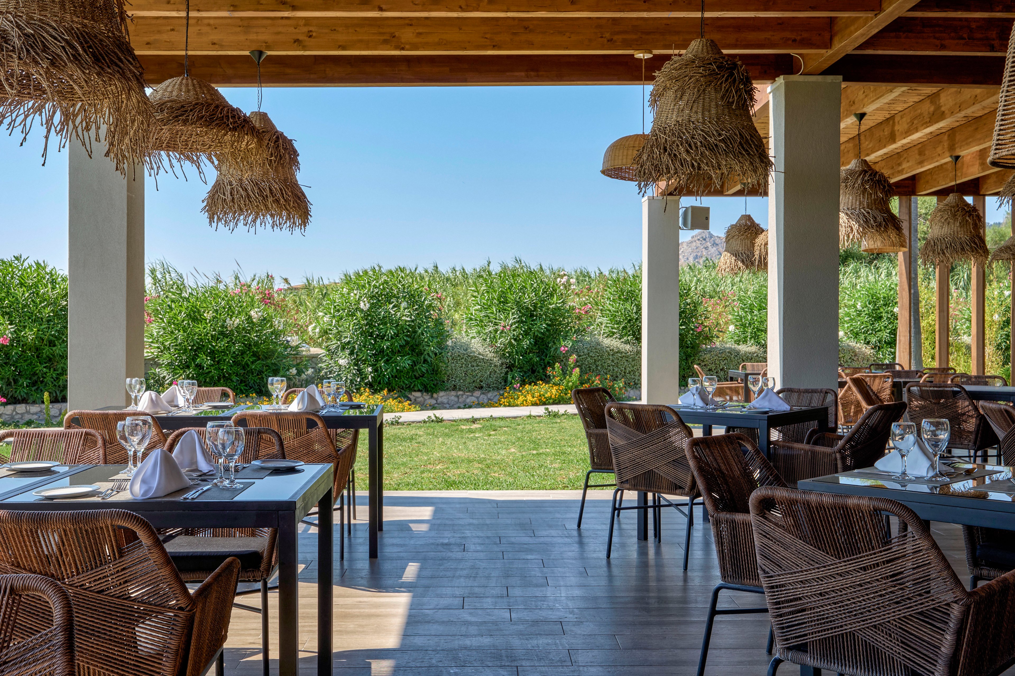 An open-air restaurant terrace features neatly arranged tables with wicker chairs and set glassware. White napkins and wine glasses are placed on dark tabletops under a wooden roof with straw-style hanging lamps. The terrace overlooks a lush green garden and hedges, with bright flowers adding color to the scene. Sunlight and clear blue sky create a relaxed, summery dining atmosphere.