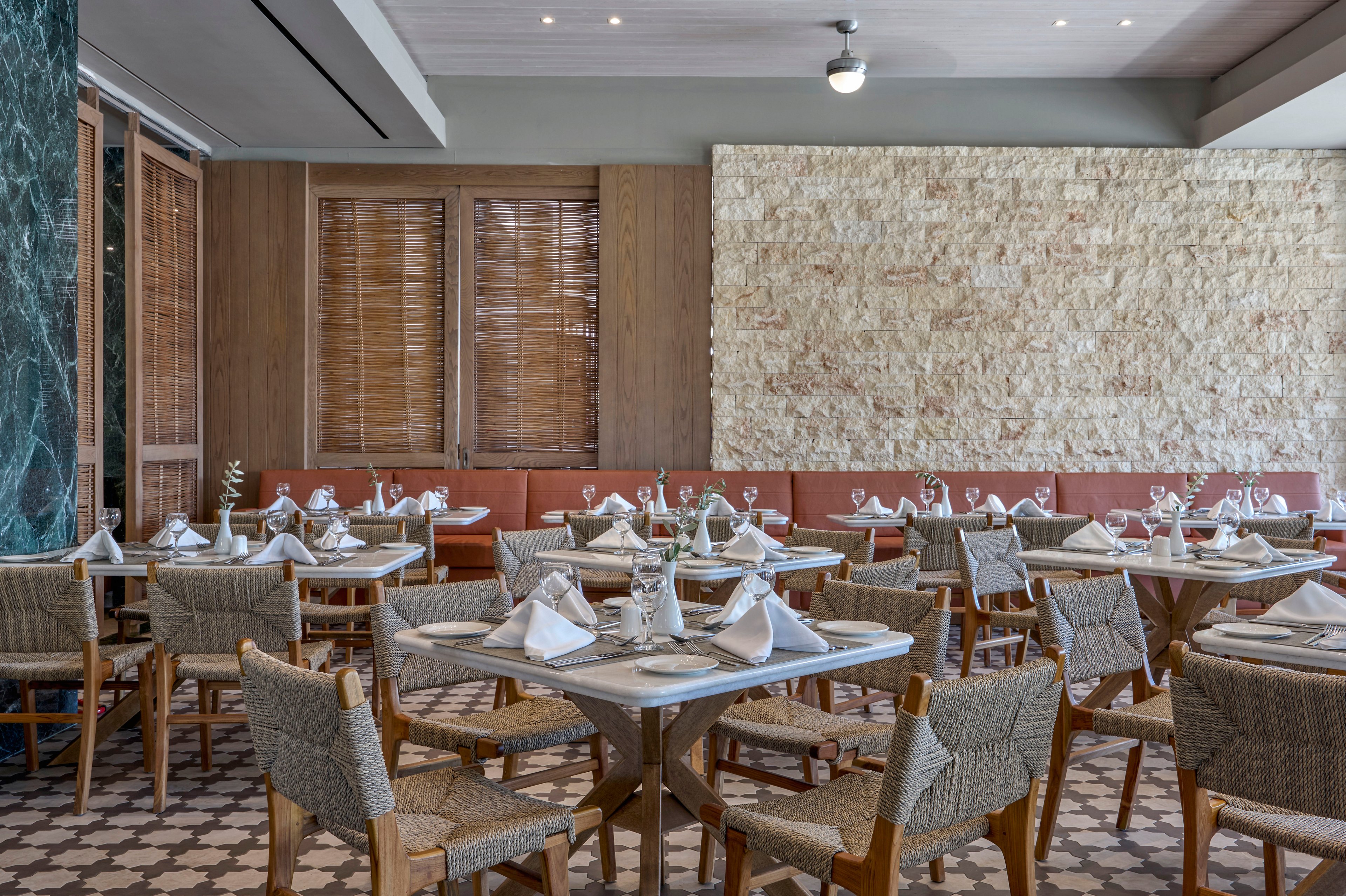 A stylish restaurant dining room is shown with neatly arranged tables and chairs. Each table is set with white napkins, glassware, and cutlery, ready for guests. The space features a patterned tile floor, cushioned bench seating, and a textured stone accent wall. Warm wood tones and soft lighting create a calm, inviting atmosphere.