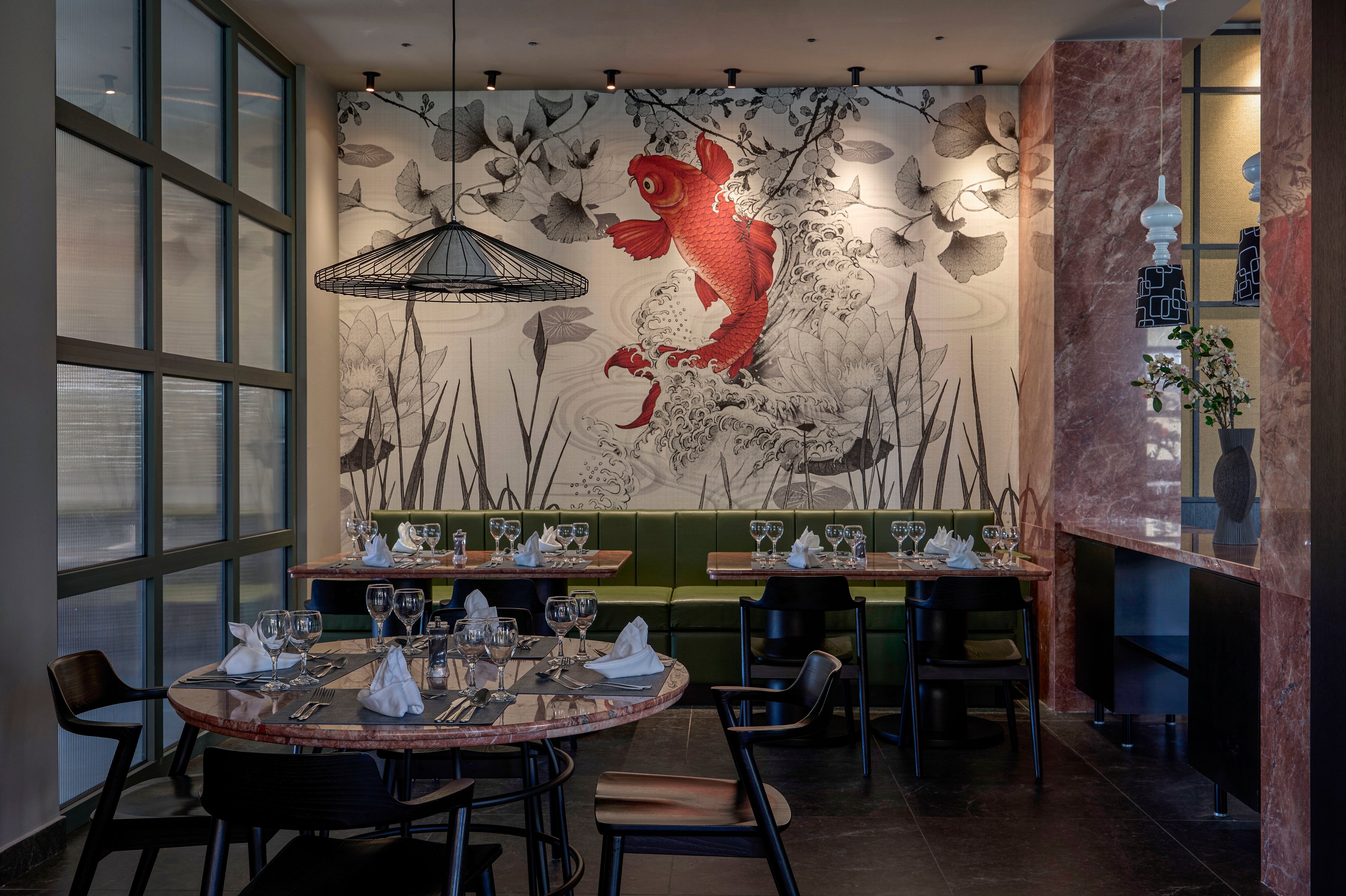 A stylish restaurant dining area features a large wall mural of a red koi fish surrounded by floral and aquatic motifs. Tables are set with wine glasses, folded napkins, and cutlery, ready for guests. Modern pendant lighting and marble columns frame the space, while green banquette seating lines the wall. The overall atmosphere is contemporary and refined with artistic decor.