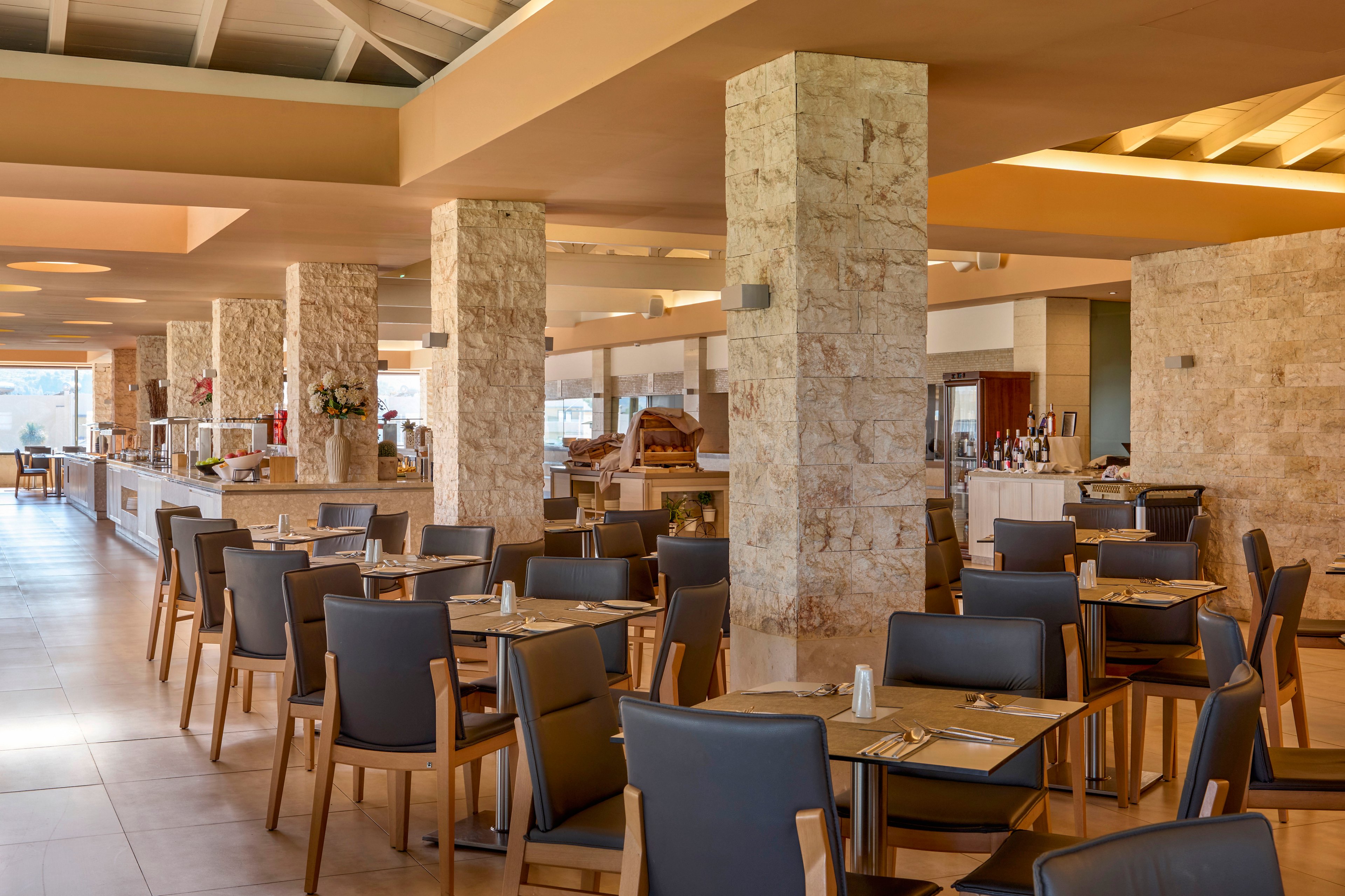 Spacious contemporary restaurant dining room with neatly arranged tables and chairs. The setting features stone-clad columns, warm neutral tones, and large windows providing natural light. Tables are set with placemats, cutlery, glasses, and condiments, suggesting readiness for guests. In the background, buffet counters and service stations indicate a hotel or resort dining experience.