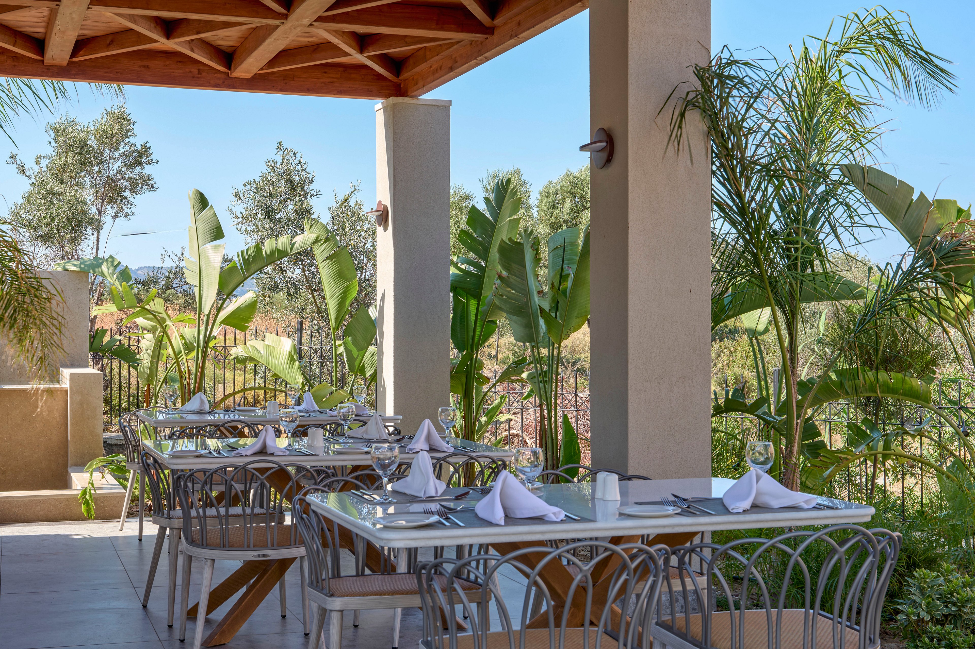 An open-air restaurant terrace features neatly arranged tables with metal chairs and white table linens. Each table is set with folded napkins, cutlery, and glassware, ready for guests. The covered patio opens onto lush tropical plants and palm trees, creating a serene garden atmosphere. Warm natural light and clear blue sky enhance the relaxed, inviting dining setting.