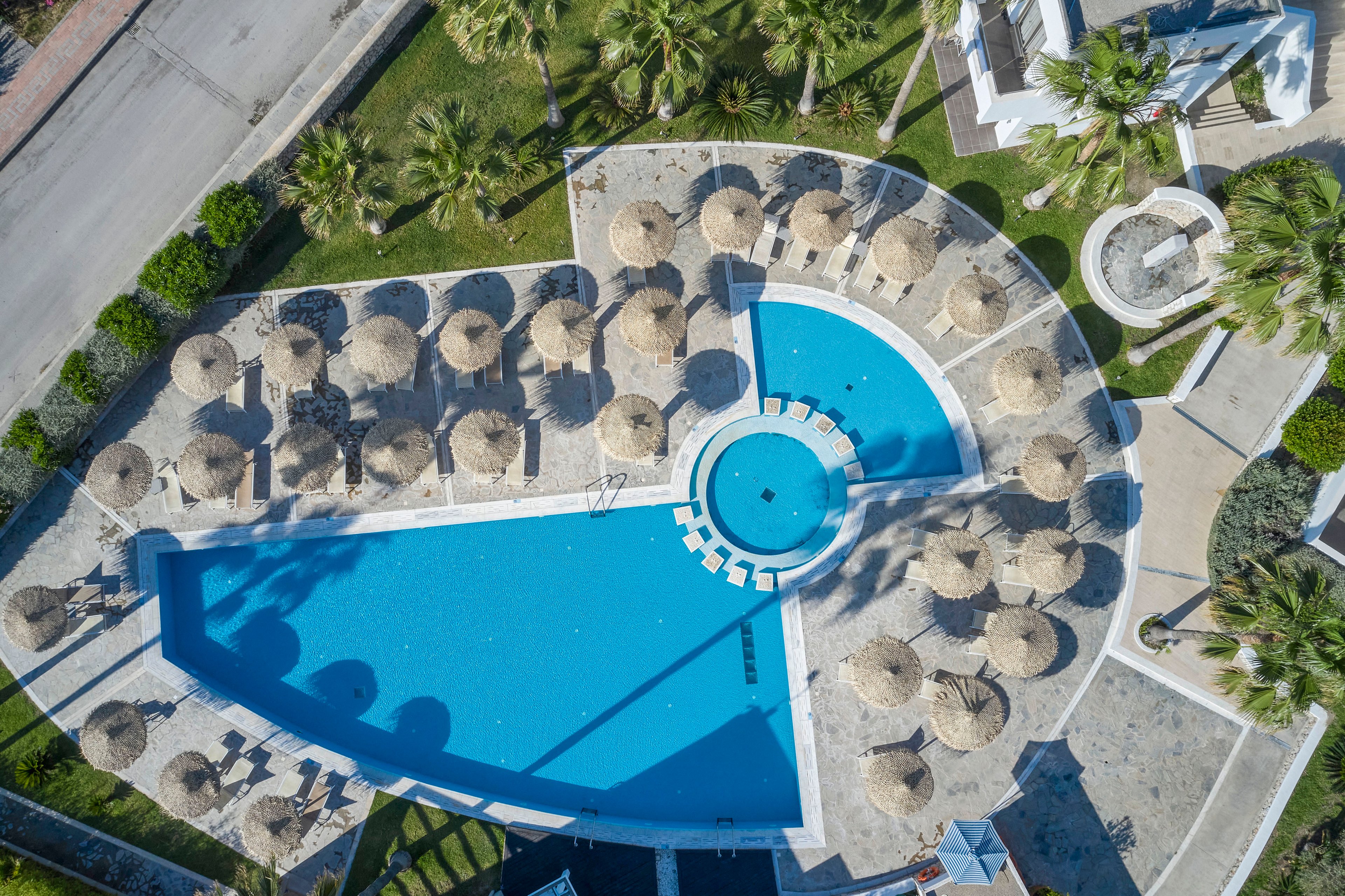A large resort swimming pool is captured from above, showing a distinctive geometric shape and a smaller circular pool connected at one end. Numerous straw parasols and sun loungers surround the pool on light stone terraces. Palm trees and landscaped greenery frame the pool area, with a road visible along the edge of the property. The bright blue water and neutral stone create a clean, inviting vacation atmosphere.