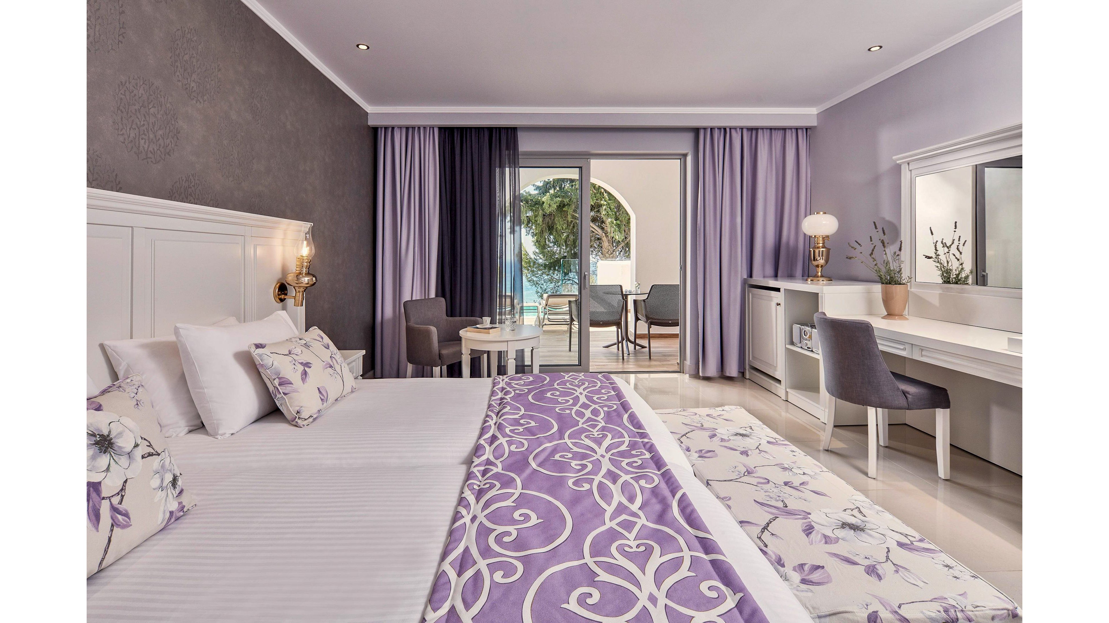 A stylish hotel room featuring a double bed adorned with purple and white floral bedding. The room includes a desk, chairs, and a balcony with a view of the sea and lush greenery. The decor showcases a soft purple palette with modern furnishings and ample natural light.