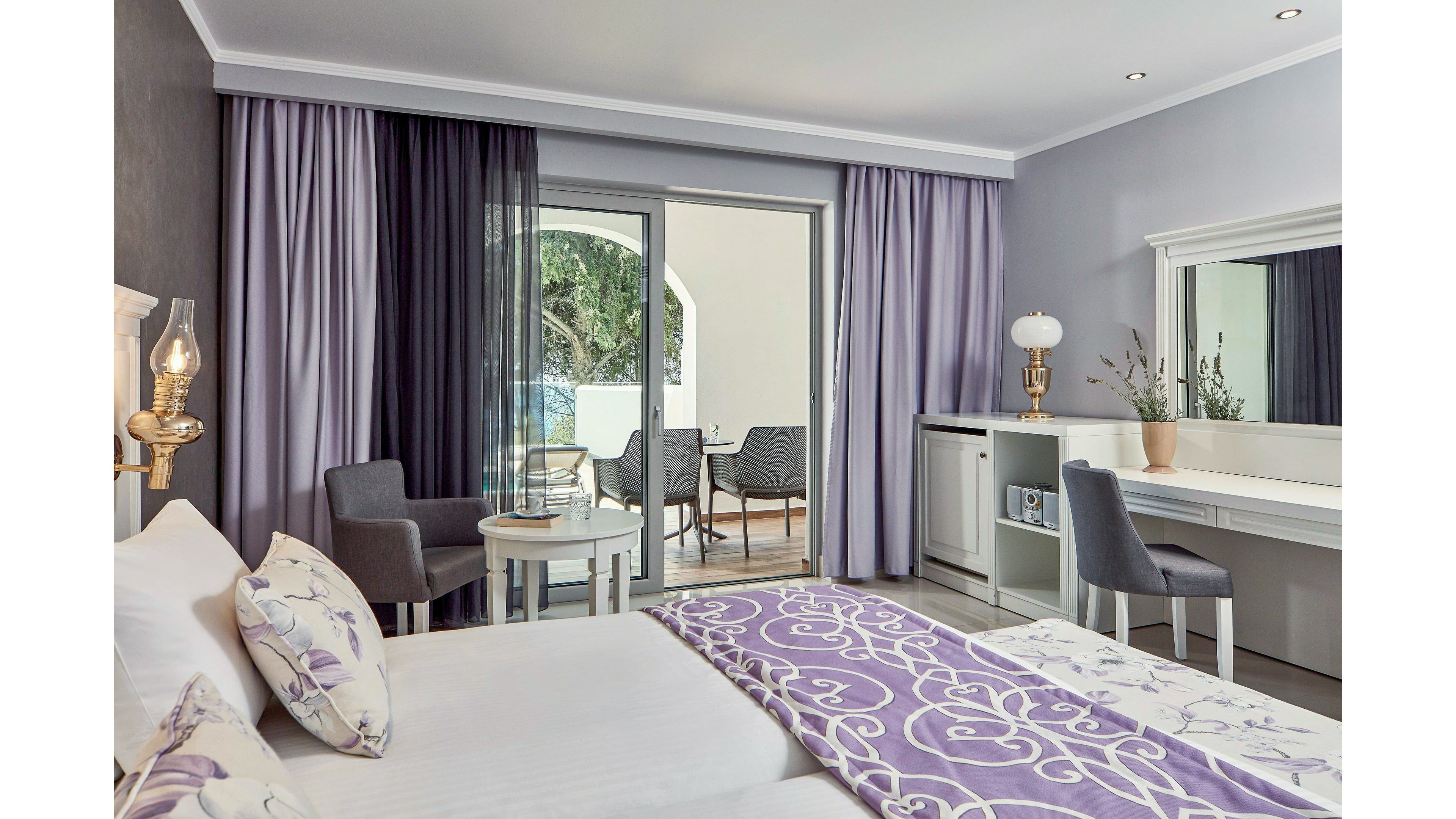 A contemporary hotel room featuring a double bed with patterned purple bedding and matching curtains. The room includes a desk, chairs, and a small table, with a balcony overlooking greenery. Neutral tones and soft lighting create a relaxing atmosphere.