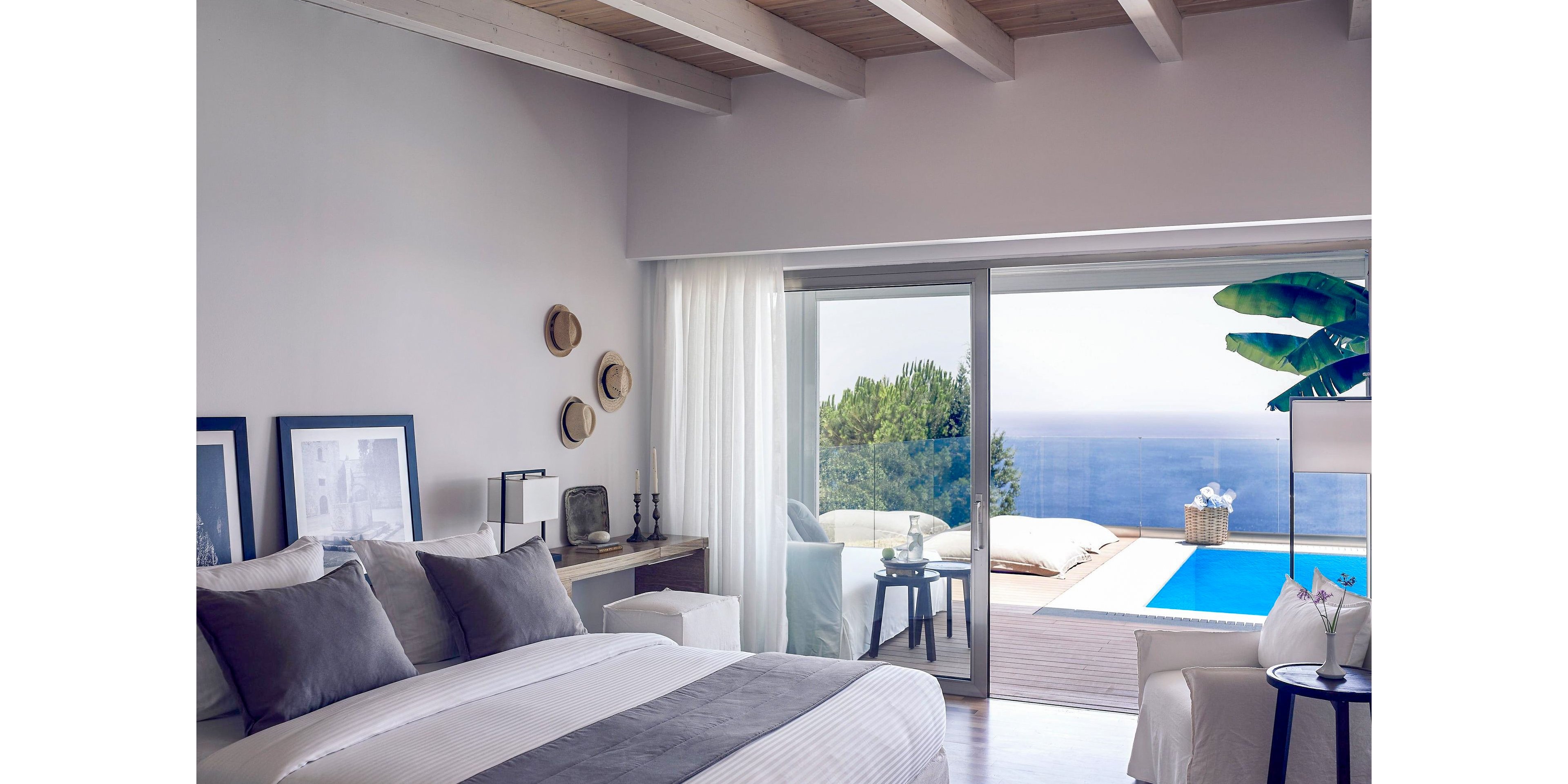 A stylish and contemporary bedroom featuring a large bed with gray and white linens. The room opens to a terrace with a private pool and stunning ocean views. Decor includes minimalist furniture, framed artwork, and natural light streaming through large glass doors.