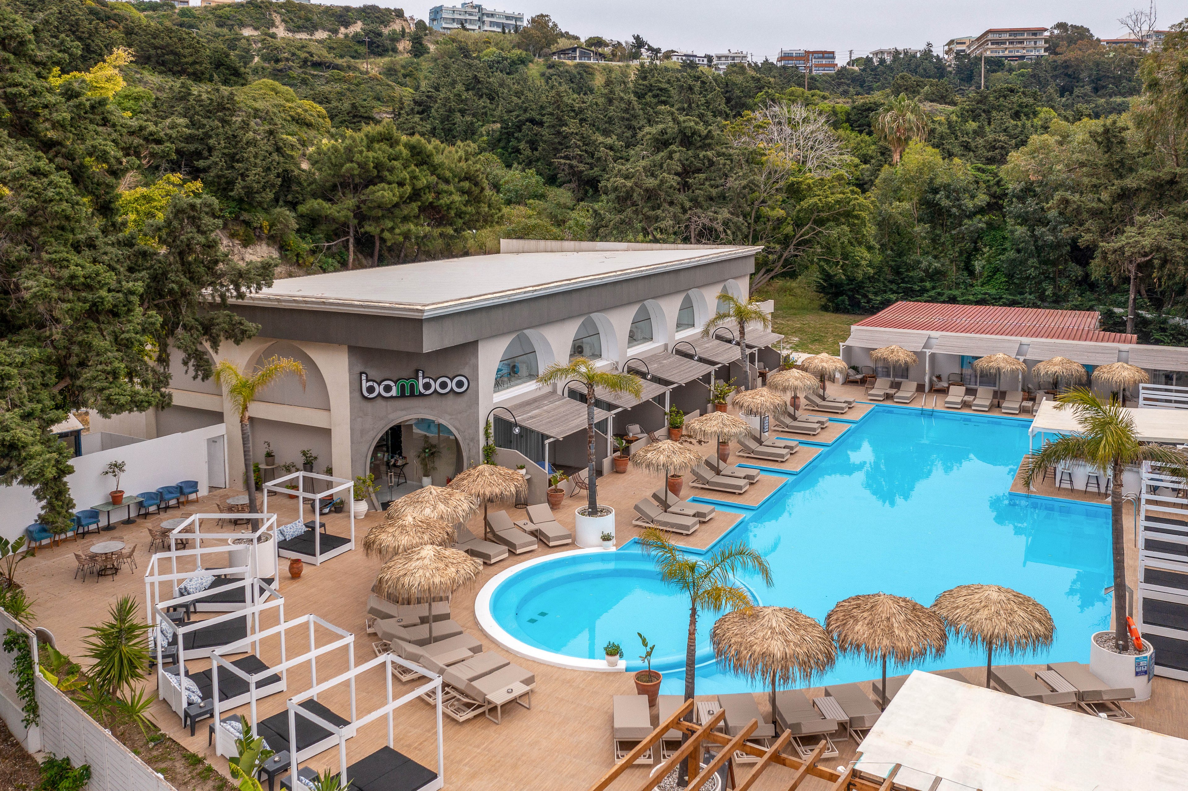 A large outdoor swimming pool is surrounded by sun loungers and straw parasols at a resort-style venue. The building beside the pool features arched openings and a sign reading "bamboo" above the entrance. White cabanas, palm trees, and neatly arranged seating create a relaxed, upscale atmosphere. Lush green hills and distant buildings frame the leisure complex in the background.