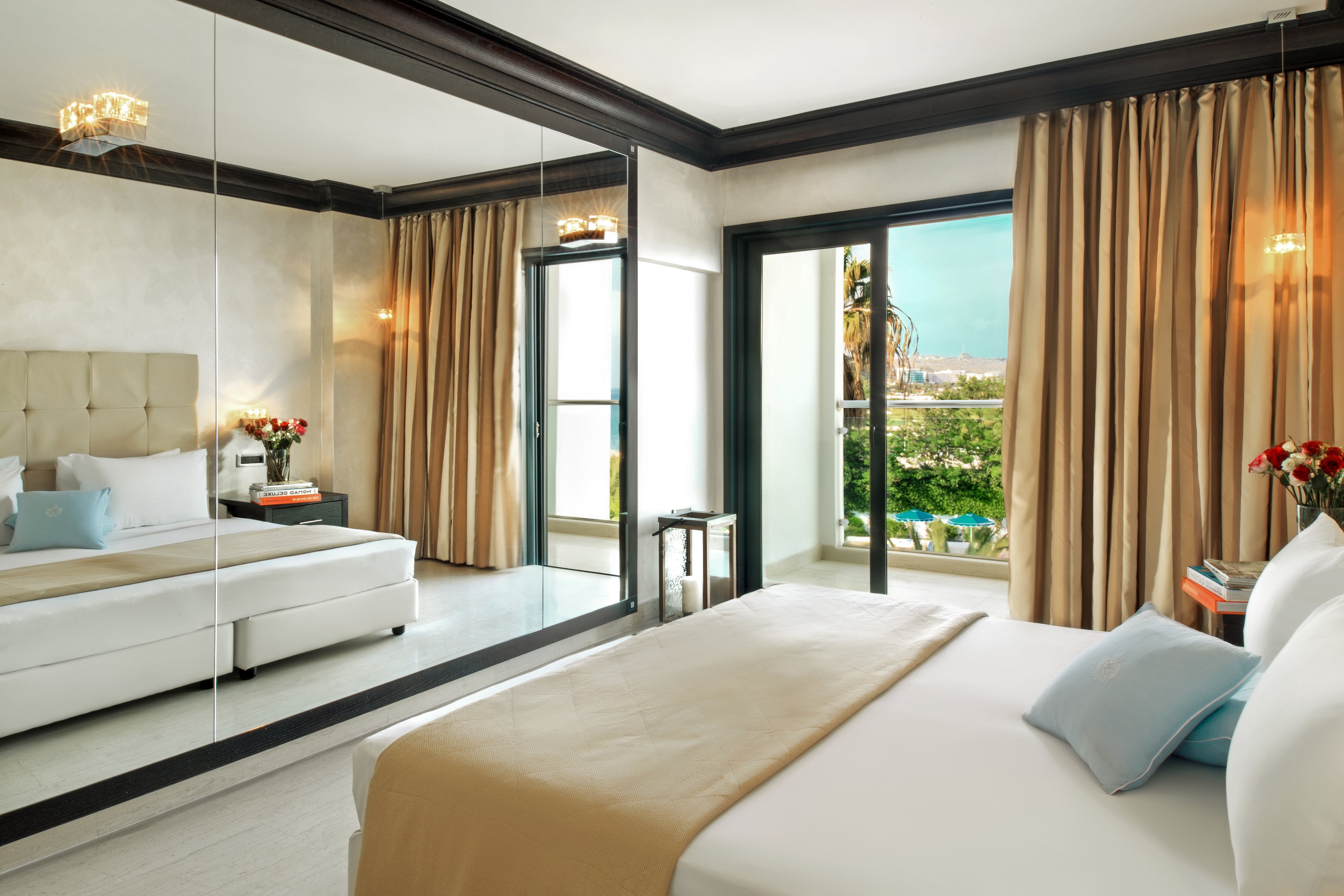 A modern and stylish hotel room featuring a large bed with beige and blue accents. The room includes a mirrored wall, bedside tables with flower arrangements, and warm lighting. The balcony offers a scenic view of greenery and a distant cityscape, creating a serene atmosphere.