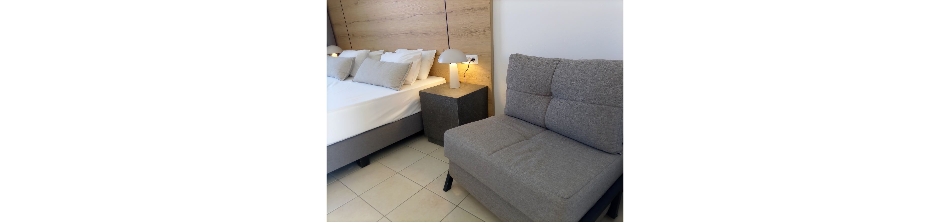 A contemporary bedroom features a neatly made bed with white linens and multiple pillows. Beside the bed is a minimalist nightstand holding a small table lamp with a rounded shade. In the foreground, a compact gray sofa or armchair sits on a light tiled floor. The room has a neutral color palette with wood paneling and soft lighting, creating a calm and inviting atmosphere.