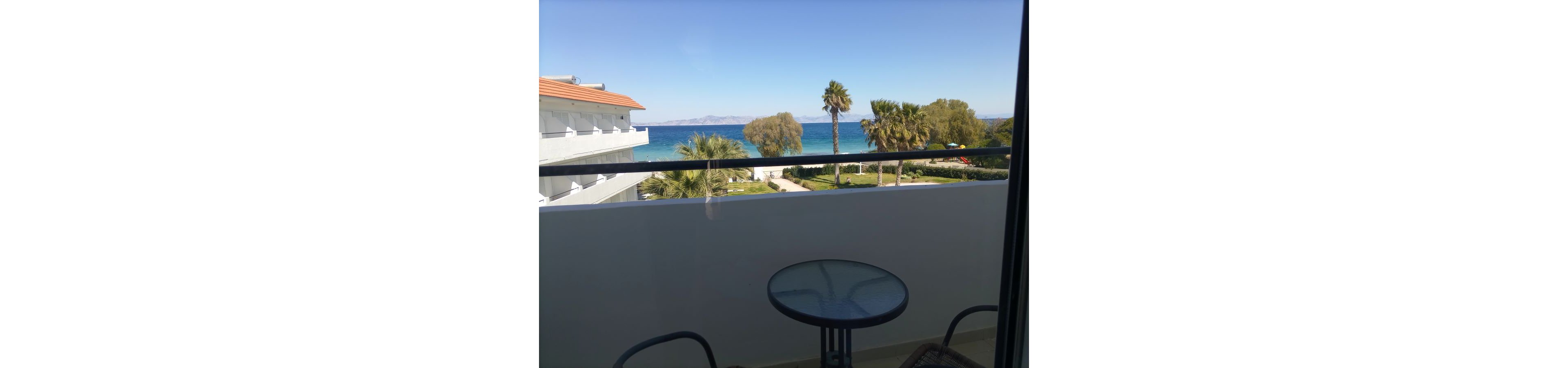 A small balcony with a round glass table and chairs overlooks a coastal resort. Beyond the balcony railing, palm trees and landscaped lawns lead toward a bright blue sea. A white multi-story building with balconies stands to the left, while distant hills are visible across the water. The clear sky and calm water create a relaxed, vacation-like atmosphere.