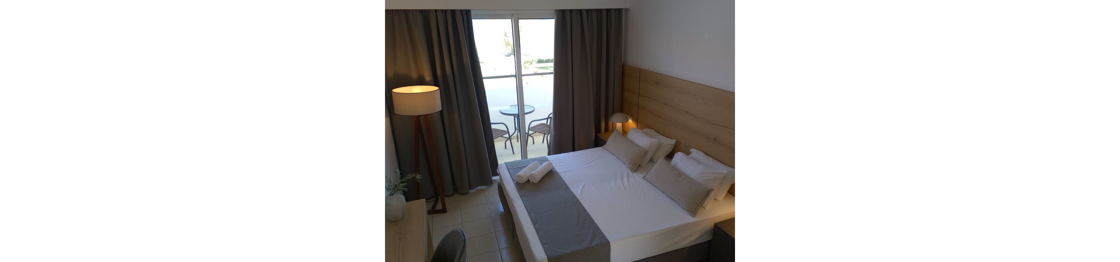 A neatly made double bed with neutral bedding is the focal point of this modern hotel room. The space features light wood furniture, tiled flooring, and soft gray curtains framing a large glass door. Outside, a small balcony with two chairs and a round table overlooks a bright exterior. Warm lighting from a floor lamp and bedside lamp creates a cozy, inviting atmosphere.