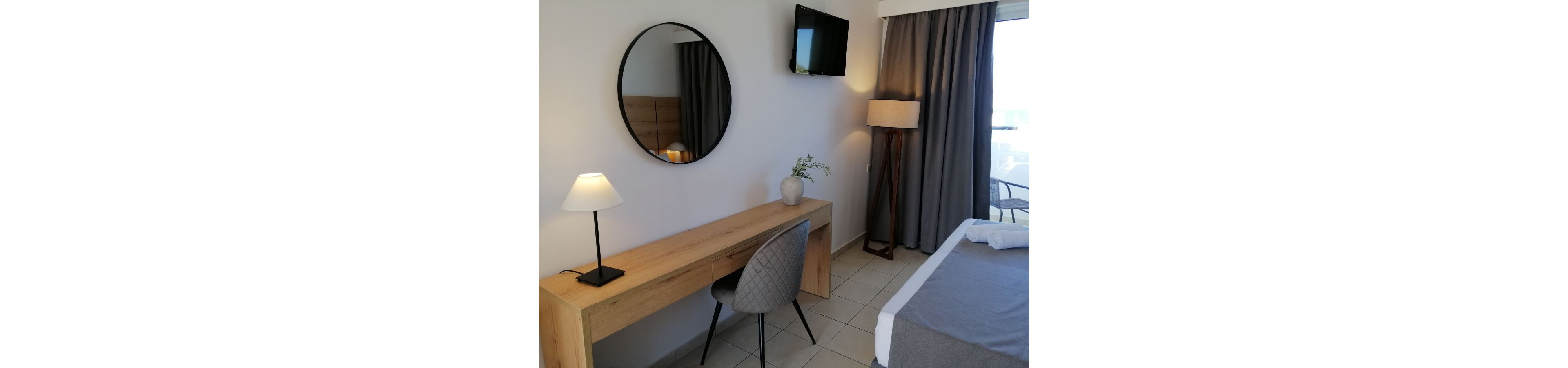 A contemporary hotel room features a wooden desk with a round wall mirror above it. A cushioned chair and a small table lamp create a simple workspace beside the bed. A floor lamp and wall-mounted television face the bed, while heavy curtains cover a window or balcony door. The neutral color palette of white, gray, and light wood gives the room a calm, minimalist atmosphere.