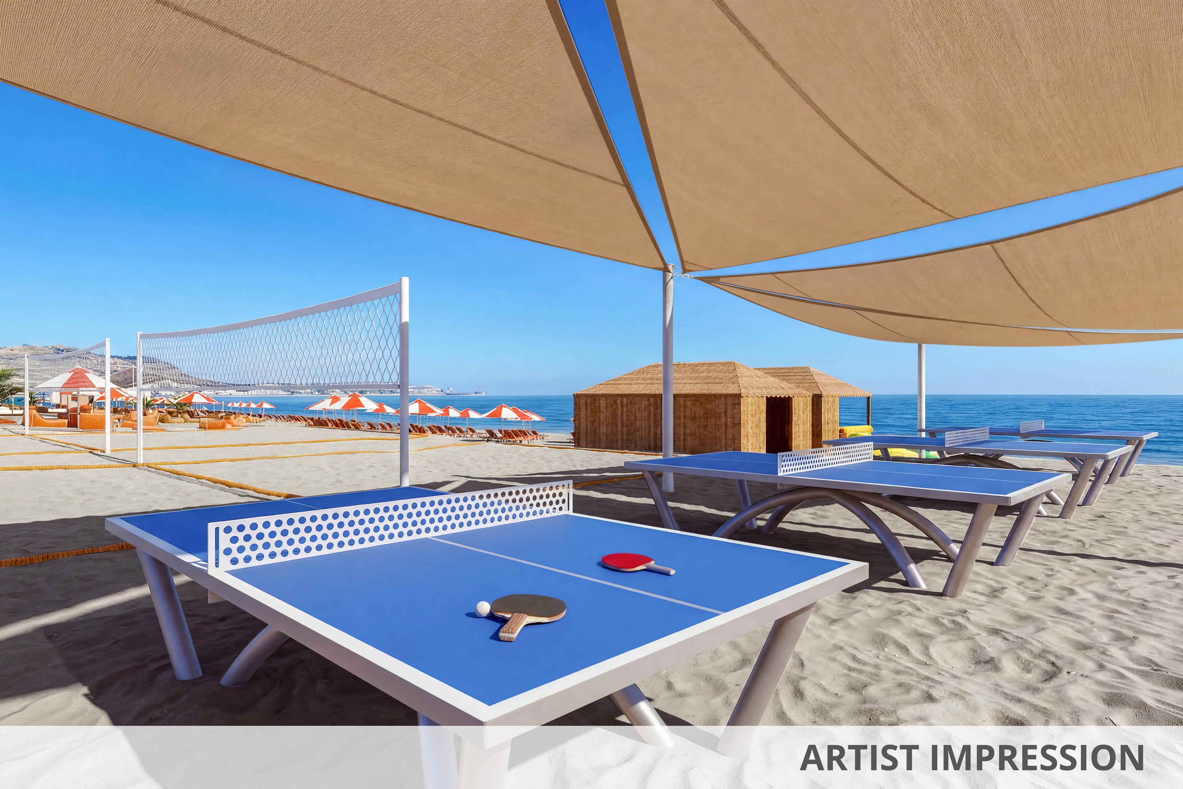 Several blue ping pong tables stand on sandy beach under large shade canopies. A red and a black paddle with a white ball rest on the nearest table, ready for a game. In the background, a beach volleyball net, orange sun loungers, and umbrellas line the shore. Calm blue sea and distant coastline complete the sunny coastal resort setting.