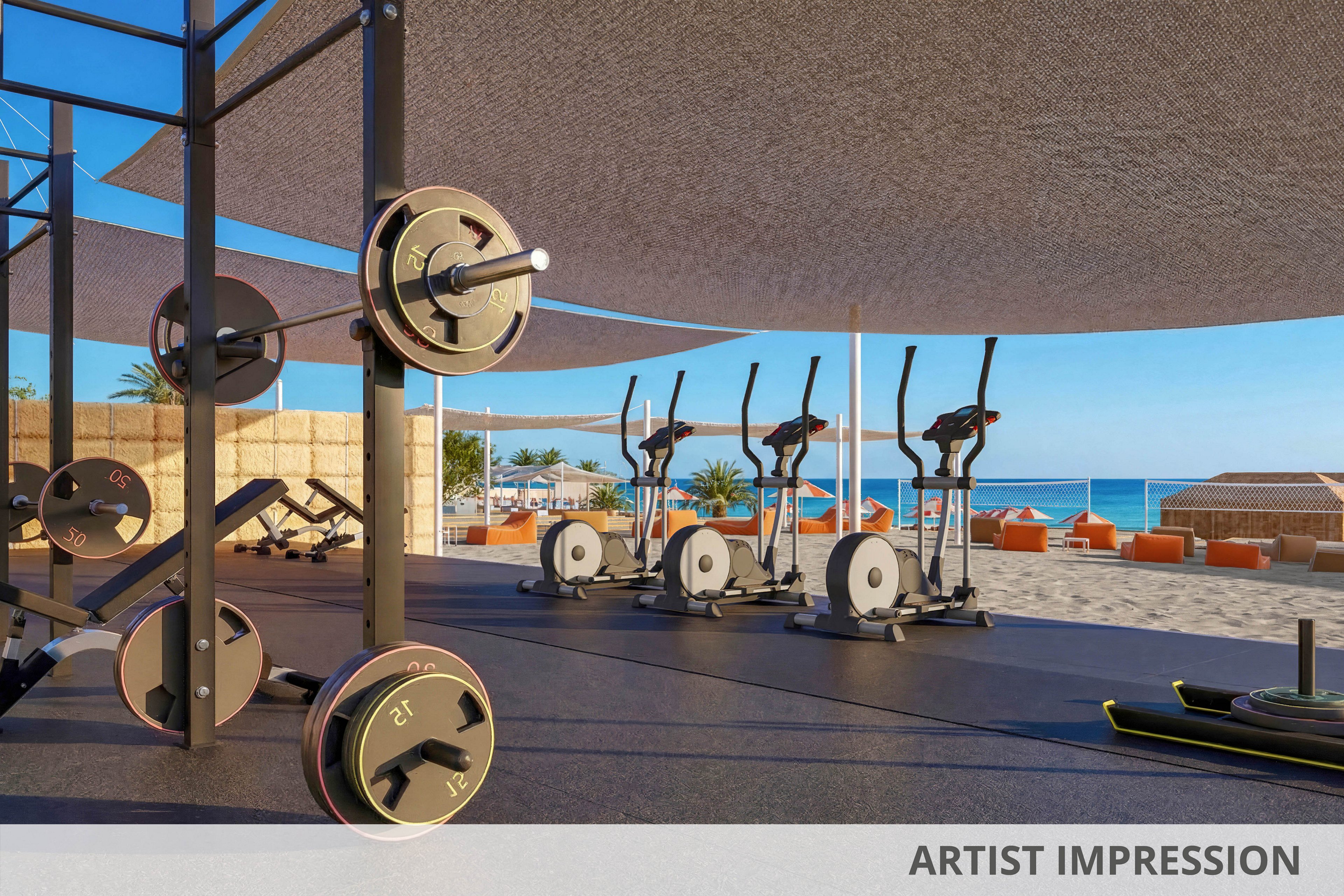 An open-air gym overlooks a sunny beachfront with clear blue water in the background. Weight racks, barbells, and stationary bikes are arranged neatly under a large shade canopy. Lounge chairs and cabanas sit near the sand, creating a resort-like atmosphere. The scene feels bright and inviting, combining fitness with a relaxing coastal setting.