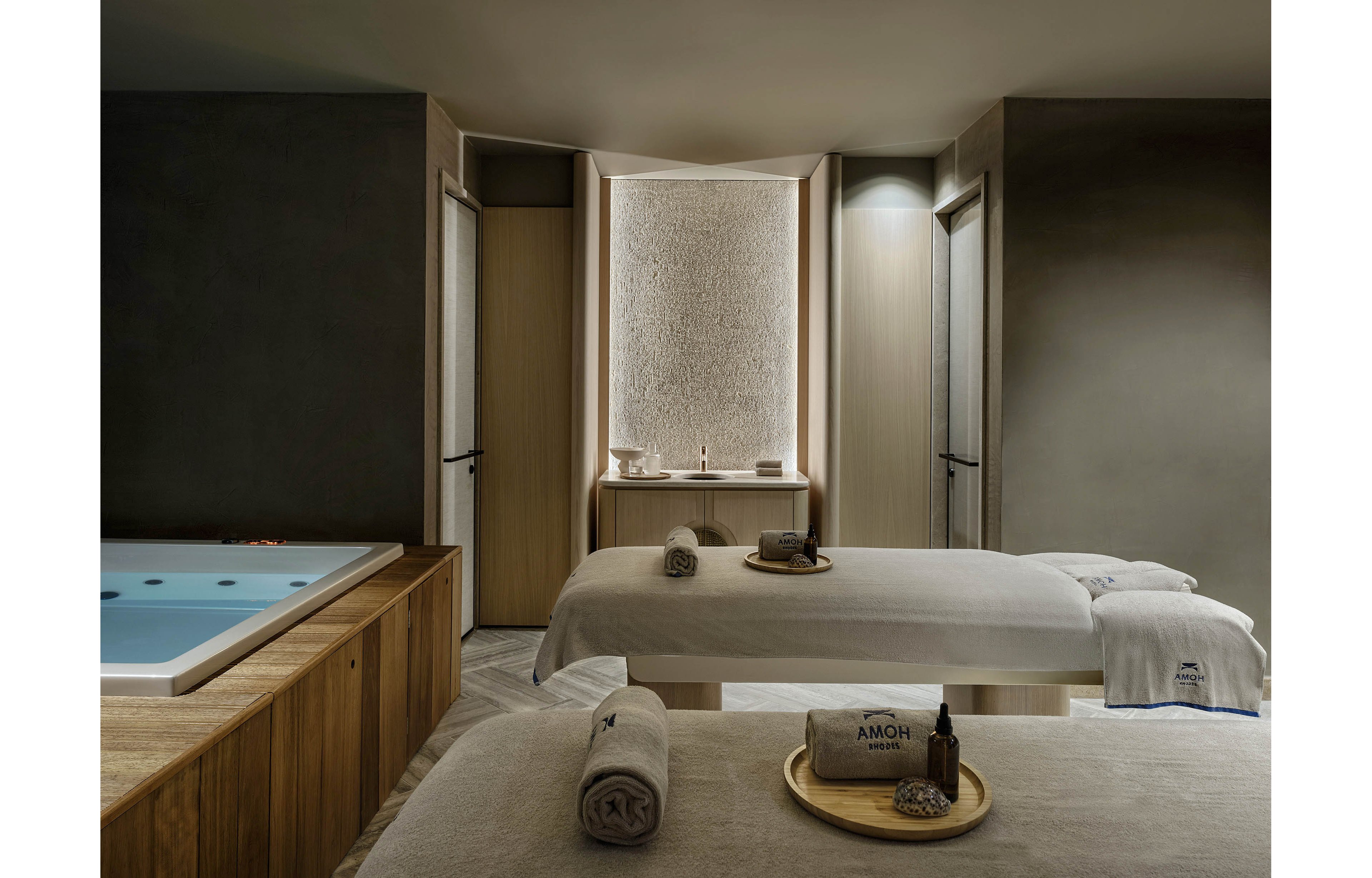 A serene spa treatment room features two massage tables neatly prepared with towels and amenities. A built-in jacuzzi tub with softly lit water sits to the side, adding to the sense of relaxation. Neutral tones, soft lighting, and natural wood surfaces create a calm and minimalist atmosphere. A tray with spa products and rolled towels is placed on one table, emphasizing wellness and comfort.
