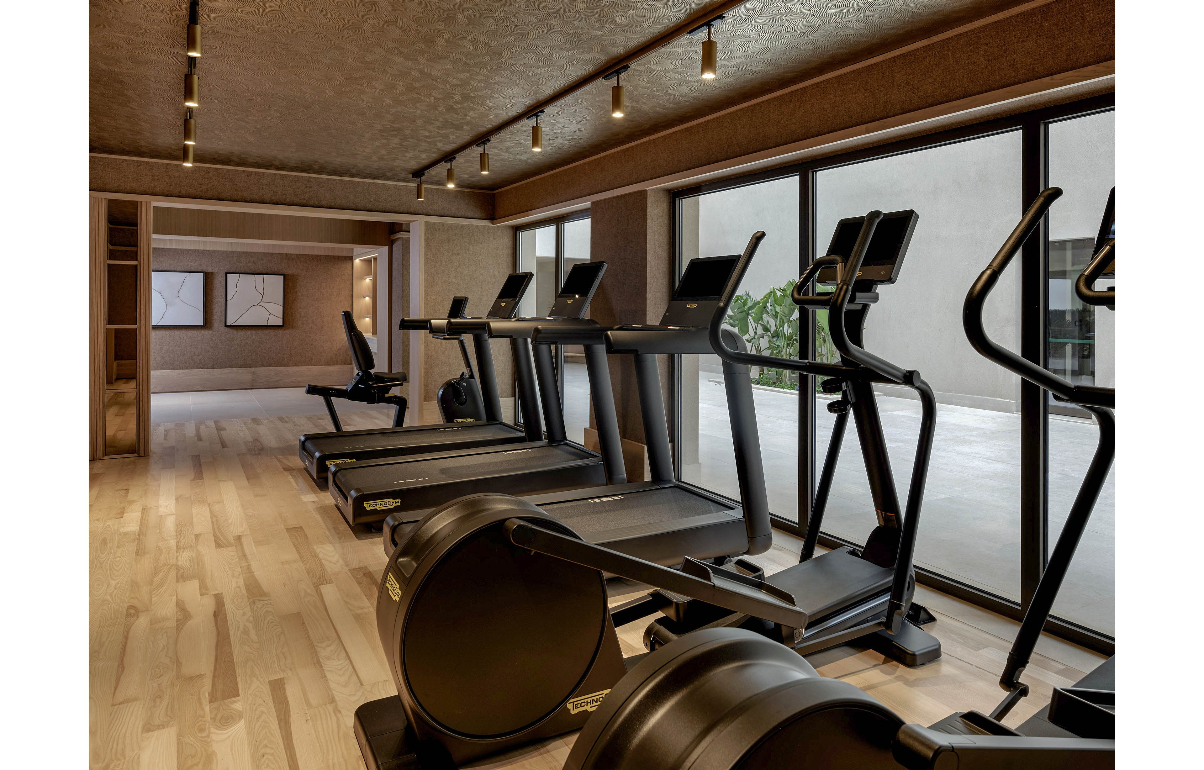 A contemporary indoor gym features several treadmills and stationary bikes neatly arranged in a row. Large floor-to-ceiling windows line one side of the room, allowing natural light to brighten the workout area. The space has warm wooden flooring, neutral-toned walls, and recessed ceiling lights that create a clean, upscale atmosphere. Framed artwork is visible in the background, adding a decorative touch to the fitness space.