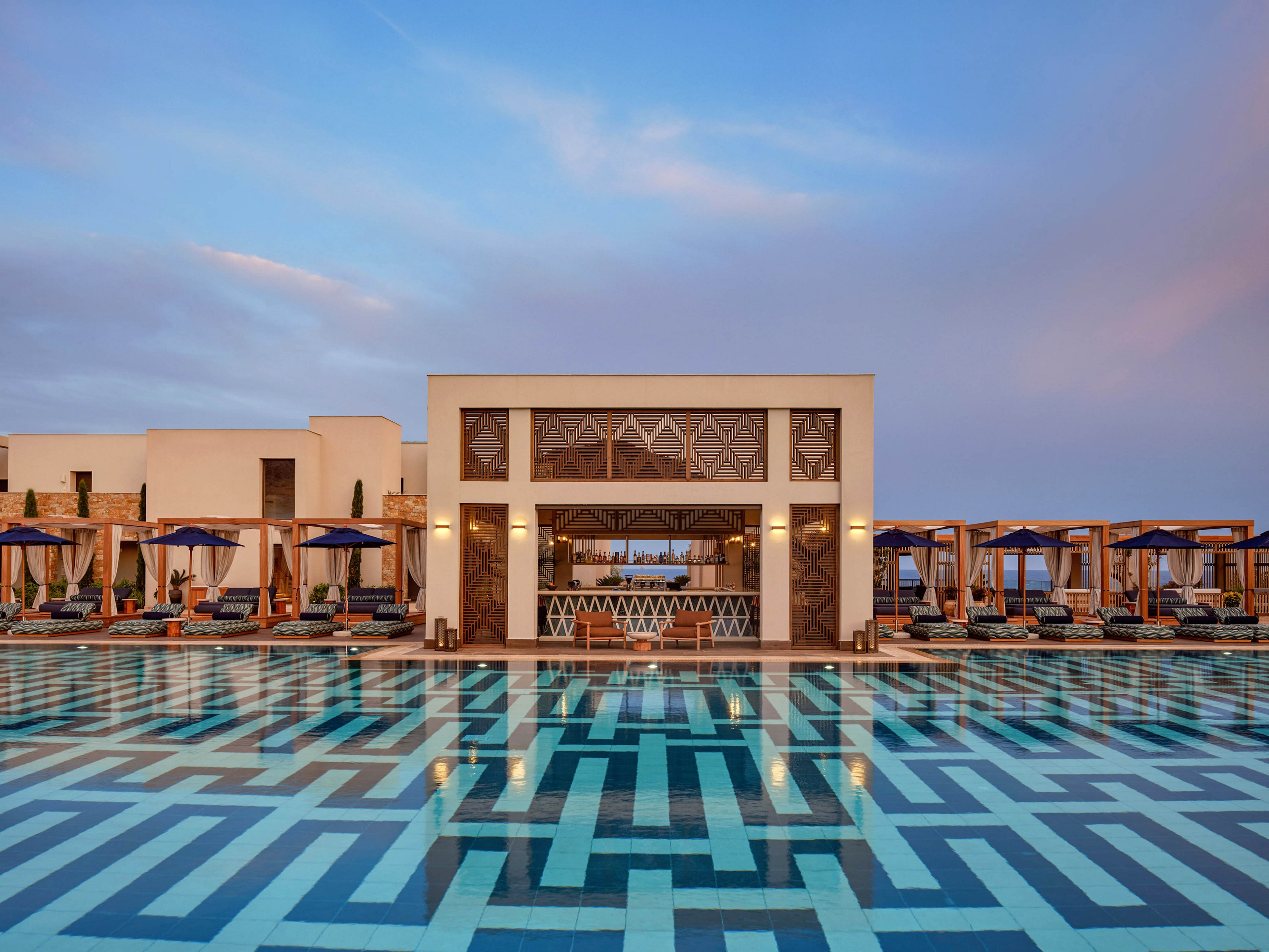 A modern resort building stands behind an expansive swimming pool with bold geometric tile patterns. Wooden cabanas and sun loungers line both sides of the pool, shaded by dark umbrellas. The architecture features clean white walls and large decorative panels above the central entrance. Soft evening light and a pastel sky create a calm, upscale vacation atmosphere.