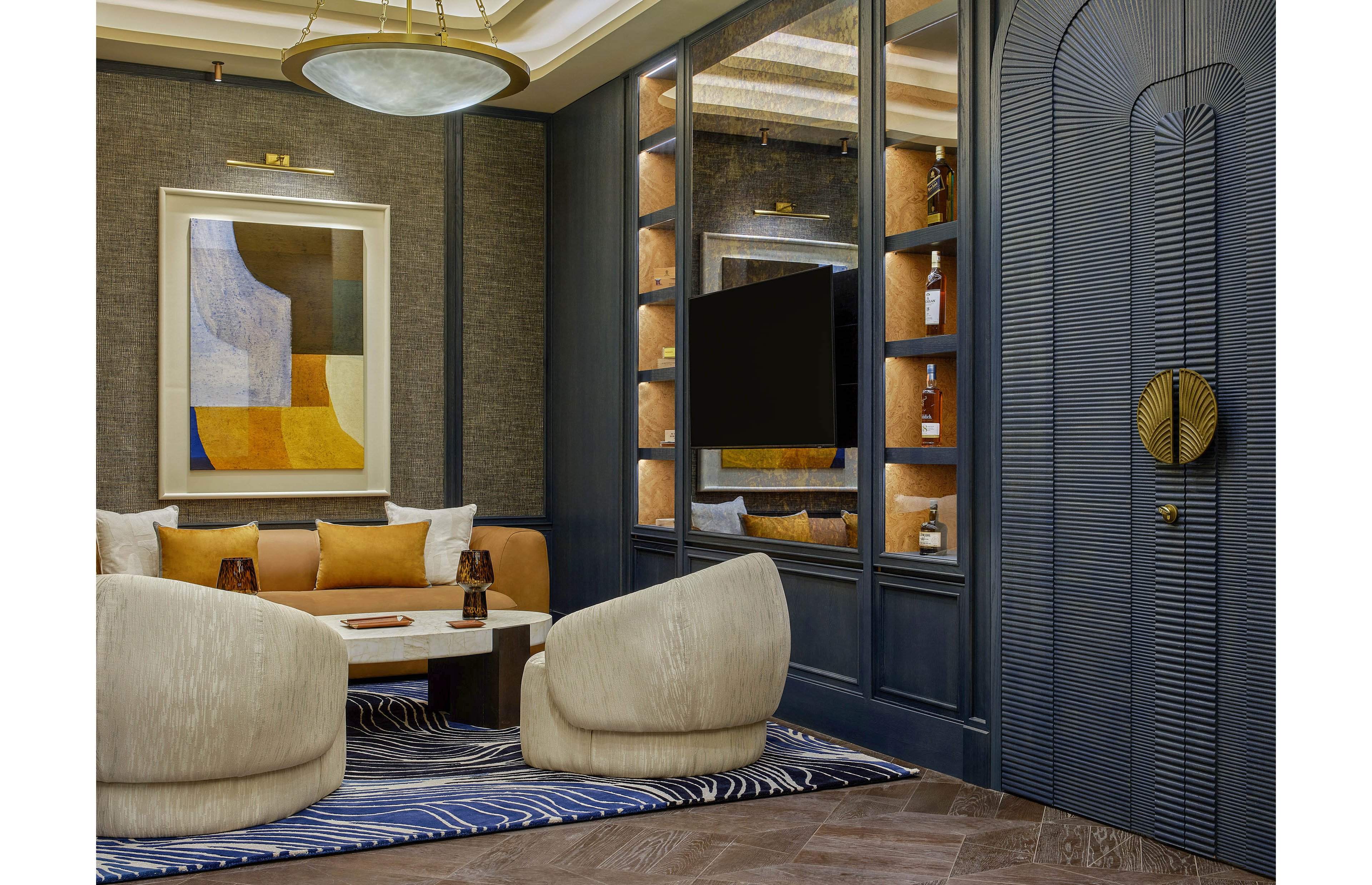 A contemporary lounge features two plush cream chairs facing a low marble coffee table. Built-in shelving with warm lighting displays bottles and decor around a wall-mounted television. A large abstract painting in muted yellow, blue, and brown tones hangs above a tan sofa with accent pillows. The space is finished with dark paneled walls, a patterned blue and white rug, and a decorative arched door with a round metallic handle.