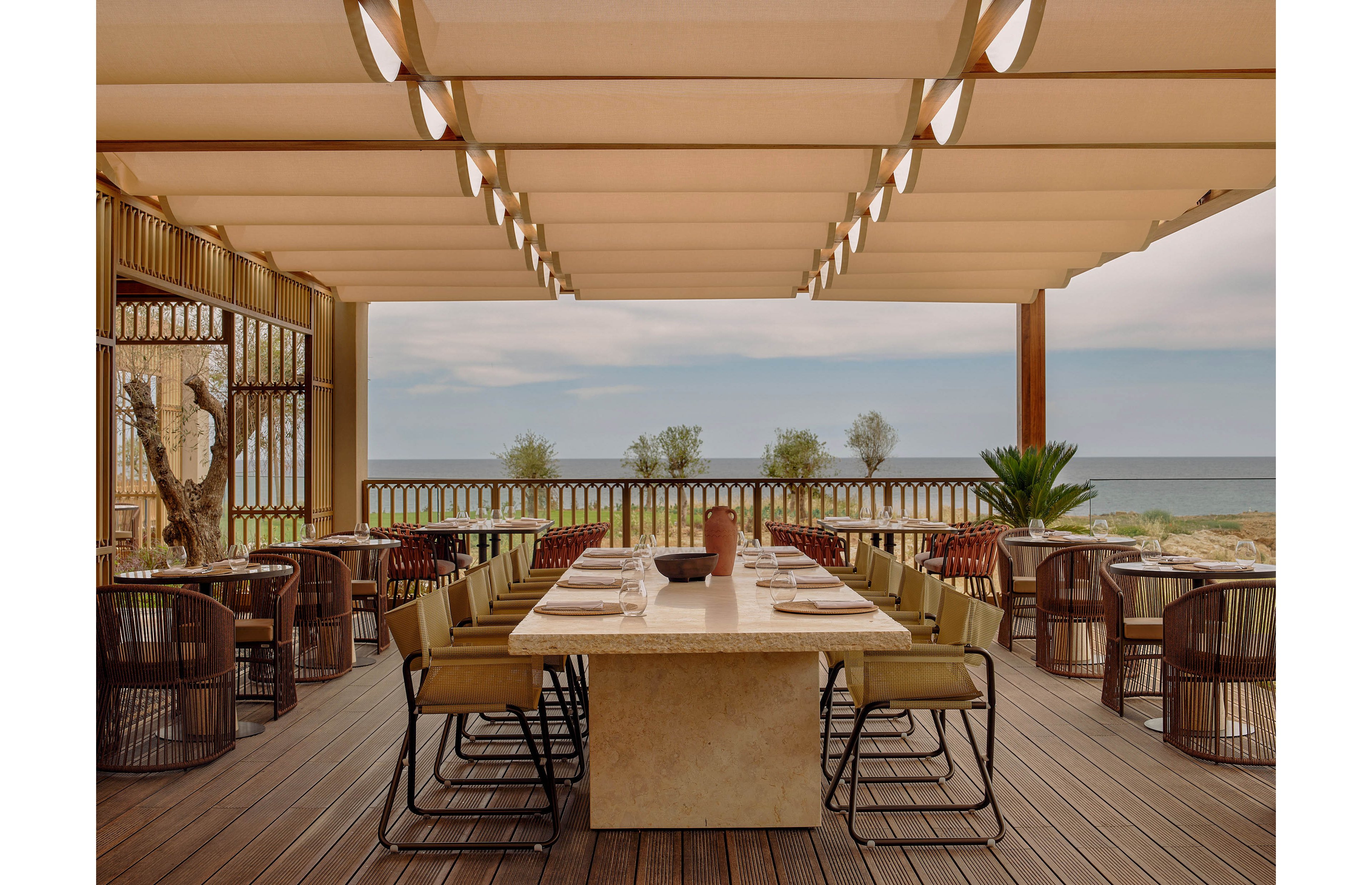 An elegant open-air restaurant features a long central dining table set with plates and glassware. The space is covered by a modern pergola that filters warm natural light onto wooden floors and contemporary chairs. Large openings frame a clear view of the ocean and sandy dunes in the background. Potted plants and natural materials create a relaxed yet upscale coastal atmosphere.