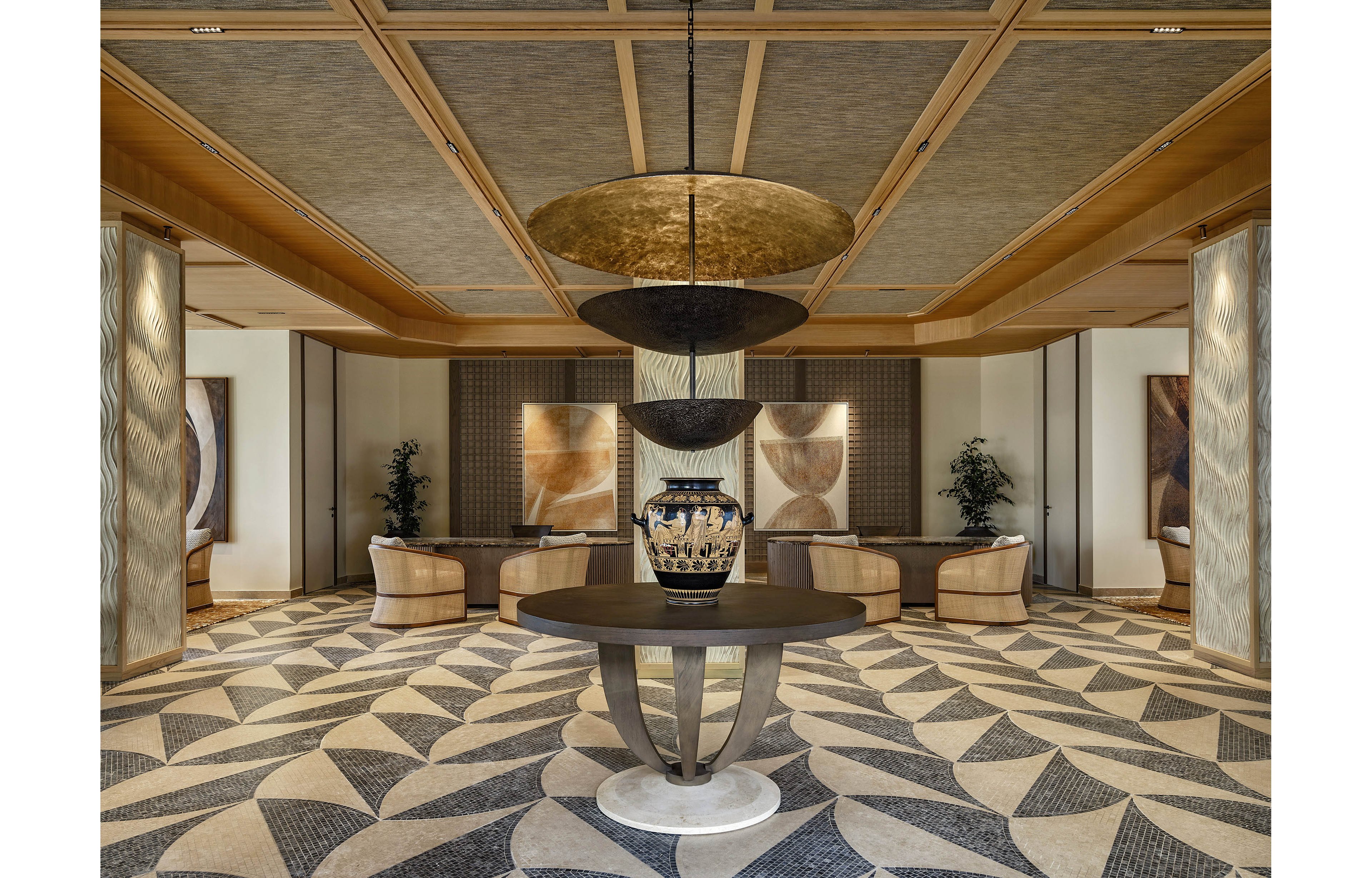 A luxurious hotel lobby features a large decorative vase displayed on a round table at the center of the room. The space is framed by curved armchairs and soft seating areas arranged symmetrically in the background. Geometric patterned flooring and layered circular ceiling fixtures create a sophisticated visual rhythm. Warm neutral tones, textured wall panels, and subtle lighting contribute to an elegant, contemporary atmosphere.