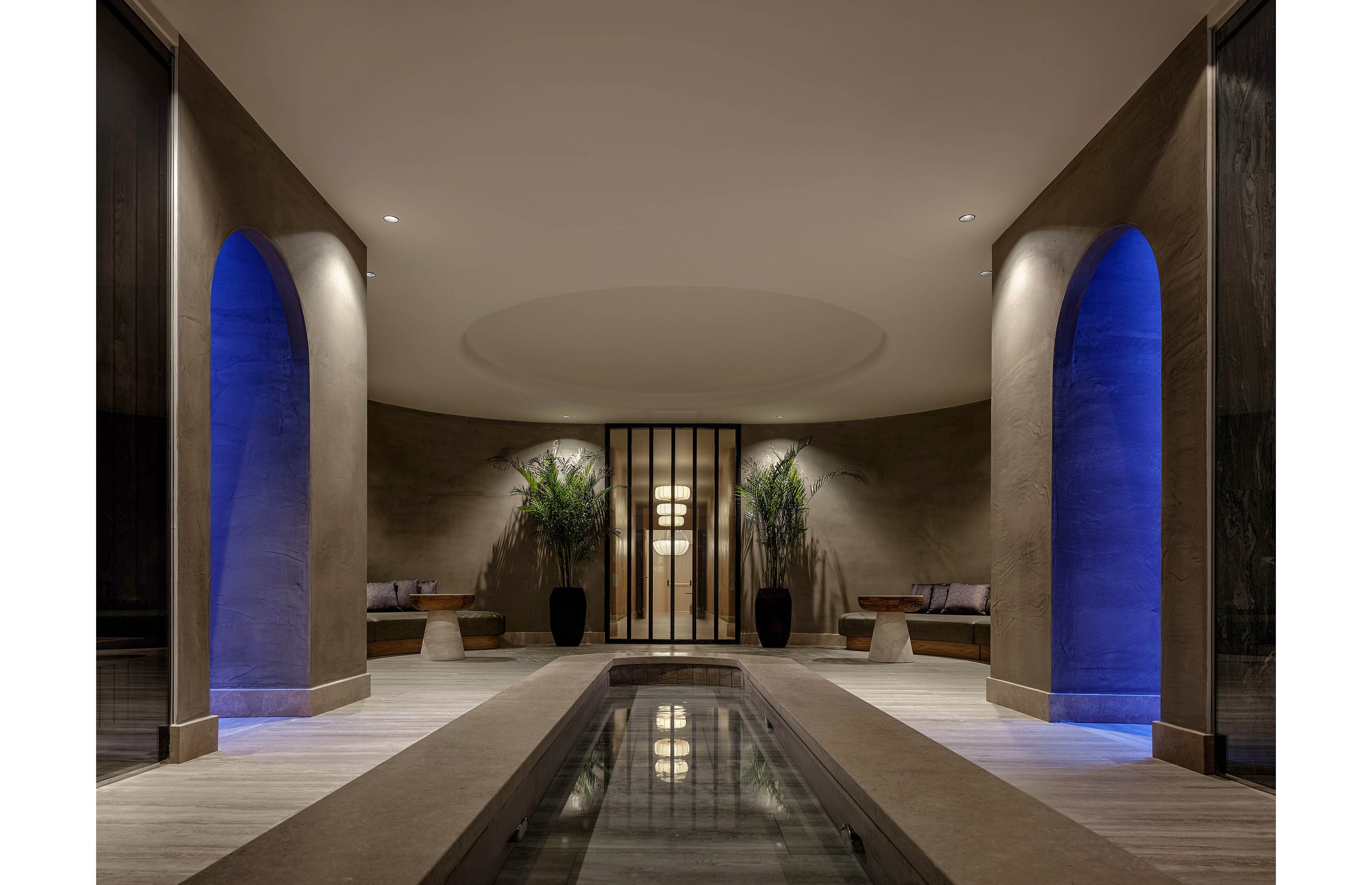 A contemporary spa corridor features a long, narrow reflective pool leading toward a softly lit seating area. Tall archways on both sides glow with blue accent lighting, framing built-in benches and side tables. Potted plants and a circular ceiling recess add organic and architectural interest to the minimalist design. The overall atmosphere is calm, luxurious, and serene, emphasizing symmetry and warm ambient light.