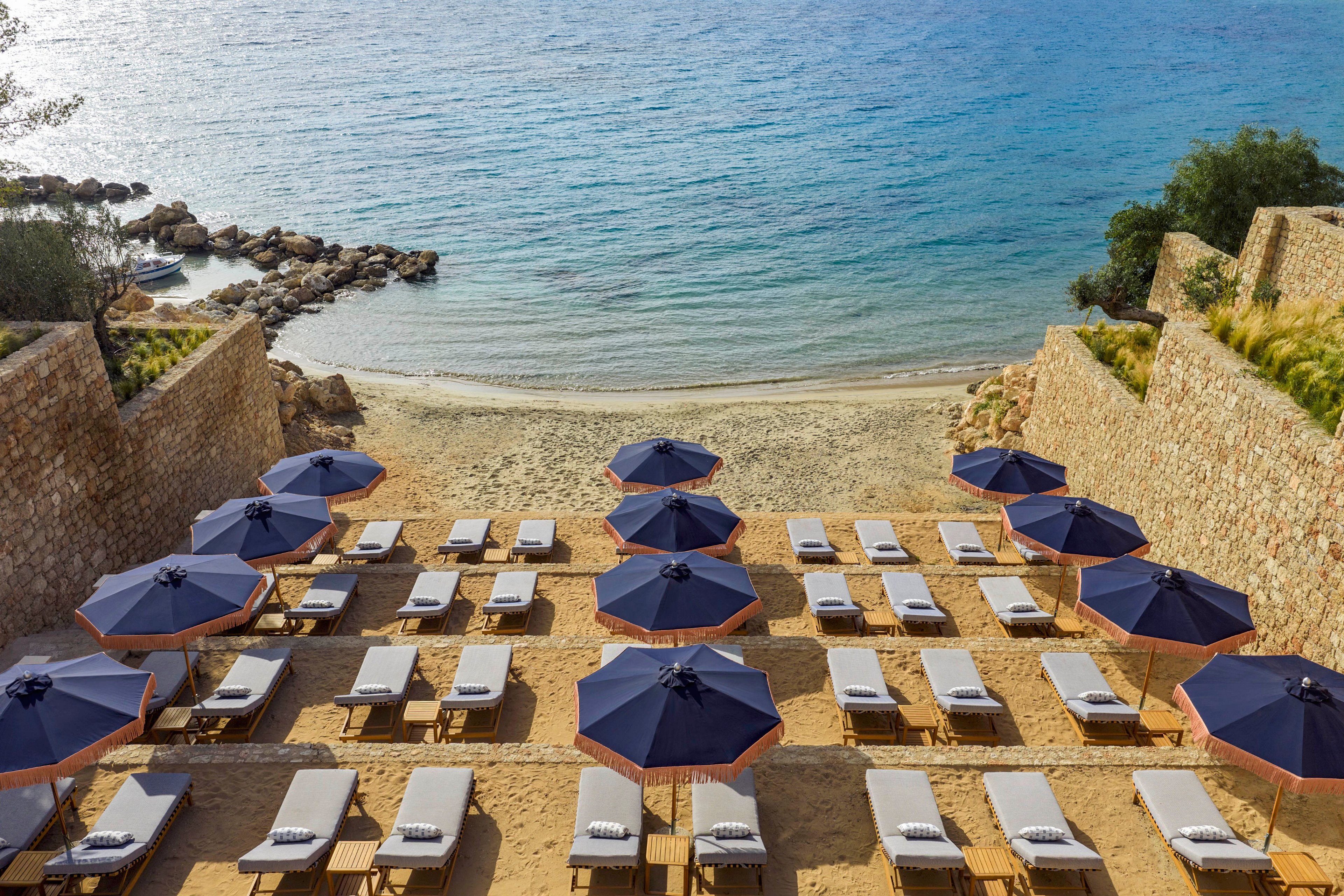 An orderly arrangement of sun loungers and dark blue umbrellas lines a sandy terrace leading down to a small beach. Stone walls frame the pathway toward the calm, clear sea in the background. The loungers are neatly organized in rows, suggesting a resort or hotel beach area prepared for guests. The scene feels peaceful and inviting, with soft natural light and gentle waves at the shoreline.