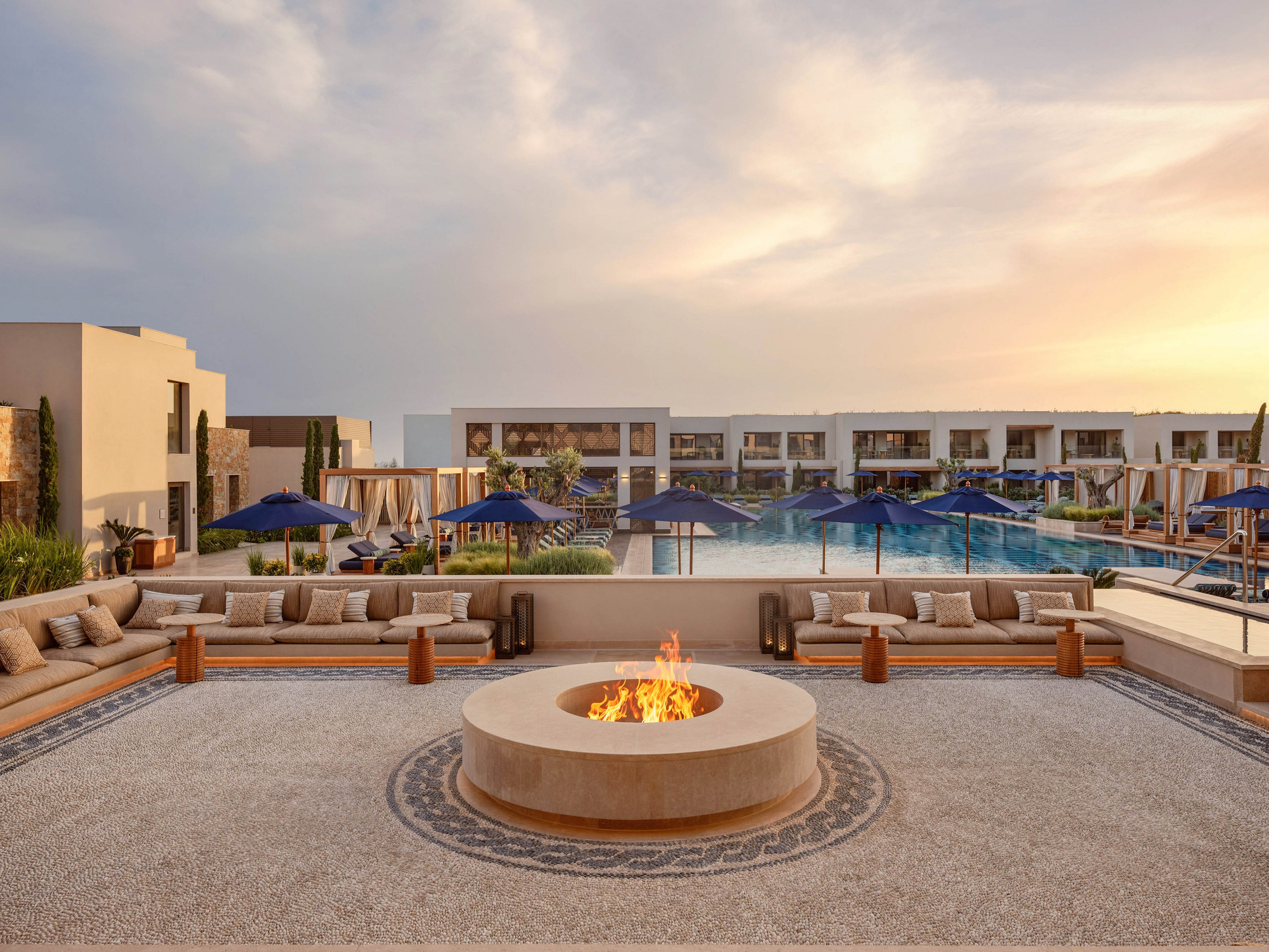 An elegant outdoor lounge features a circular fire pit surrounded by cushioned seating. Beyond the seating area, a large resort-style swimming pool is lined with blue umbrellas and cabanas. Modern low-rise buildings frame the background under a soft, pastel evening sky. The scene conveys a tranquil, upscale vacation atmosphere with warm and cool tones in balance.
