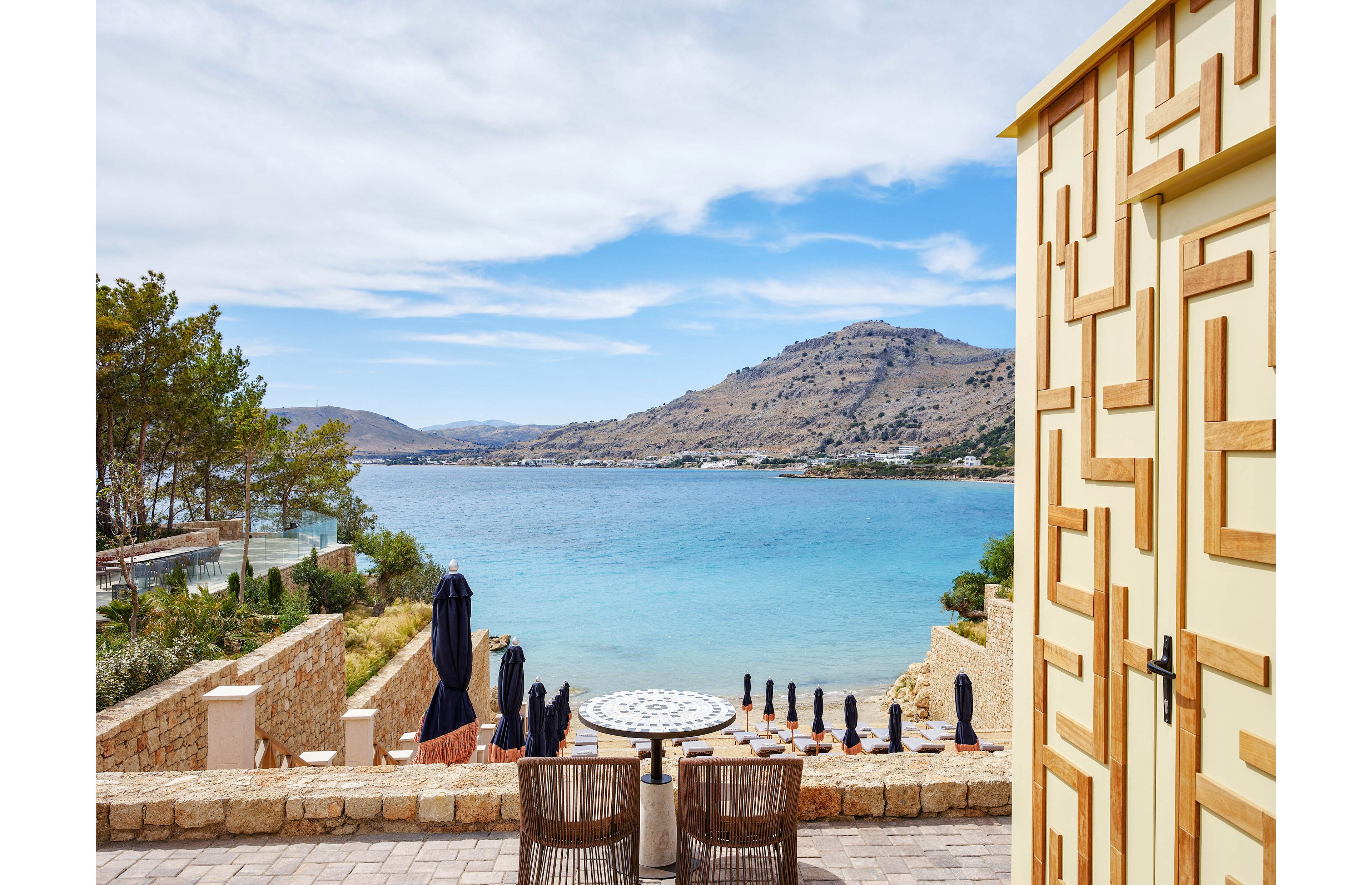 An outdoor restaurant terrace overlooks a calm blue bay framed by rocky hills. A round table with patterned top and neatly arranged place settings sits in the foreground, with wicker chairs facing the water. Stone walls, steps, and a geometric wooden structure add architectural interest to the scene. The bright daylight and soft clouds create a relaxed, inviting coastal atmosphere.