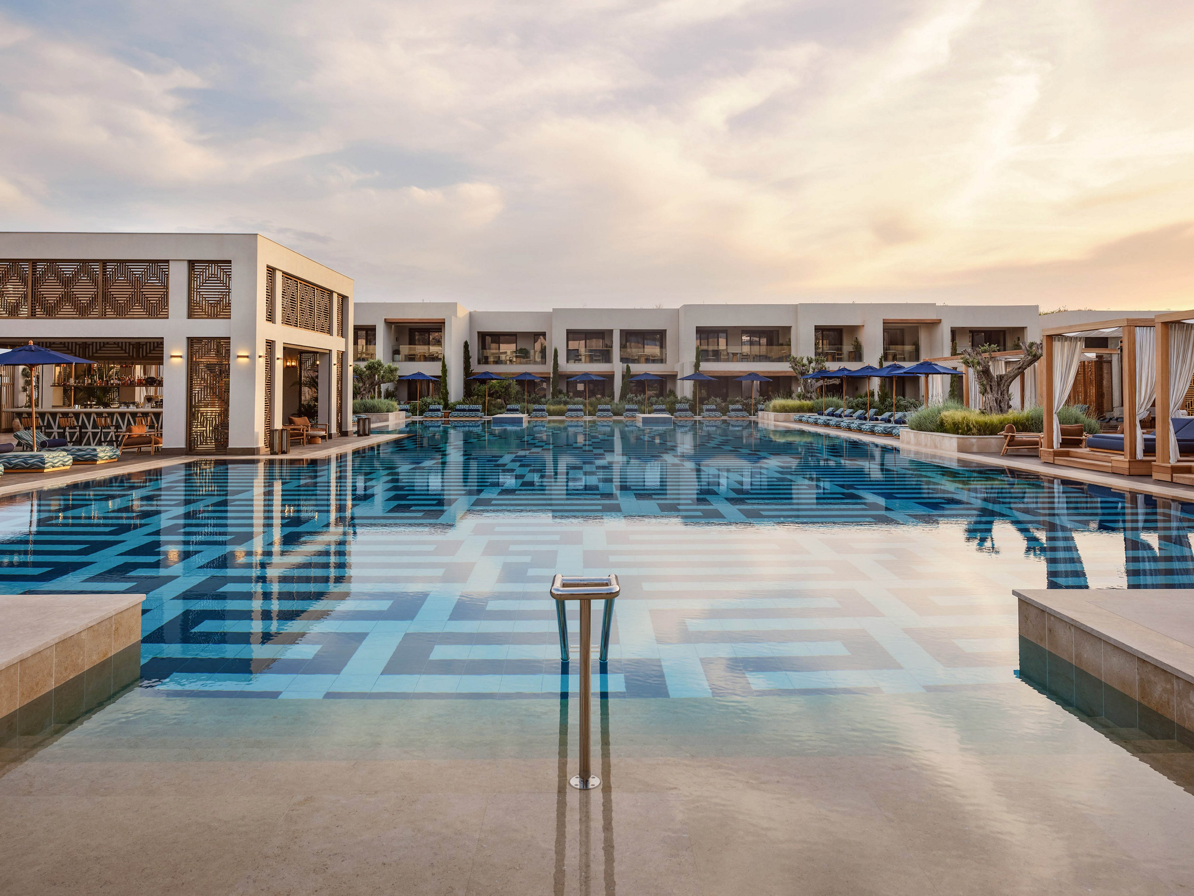 A large, tranquil infinity pool stretches toward a row of modern resort suites. The water reflects the soft evening sky and geometric tile patterns on the pool floor. Sun loungers and cabanas with blue umbrellas line the sides, suggesting a relaxing vacation setting. The overall scene feels calm, upscale, and inviting for leisure travelers.