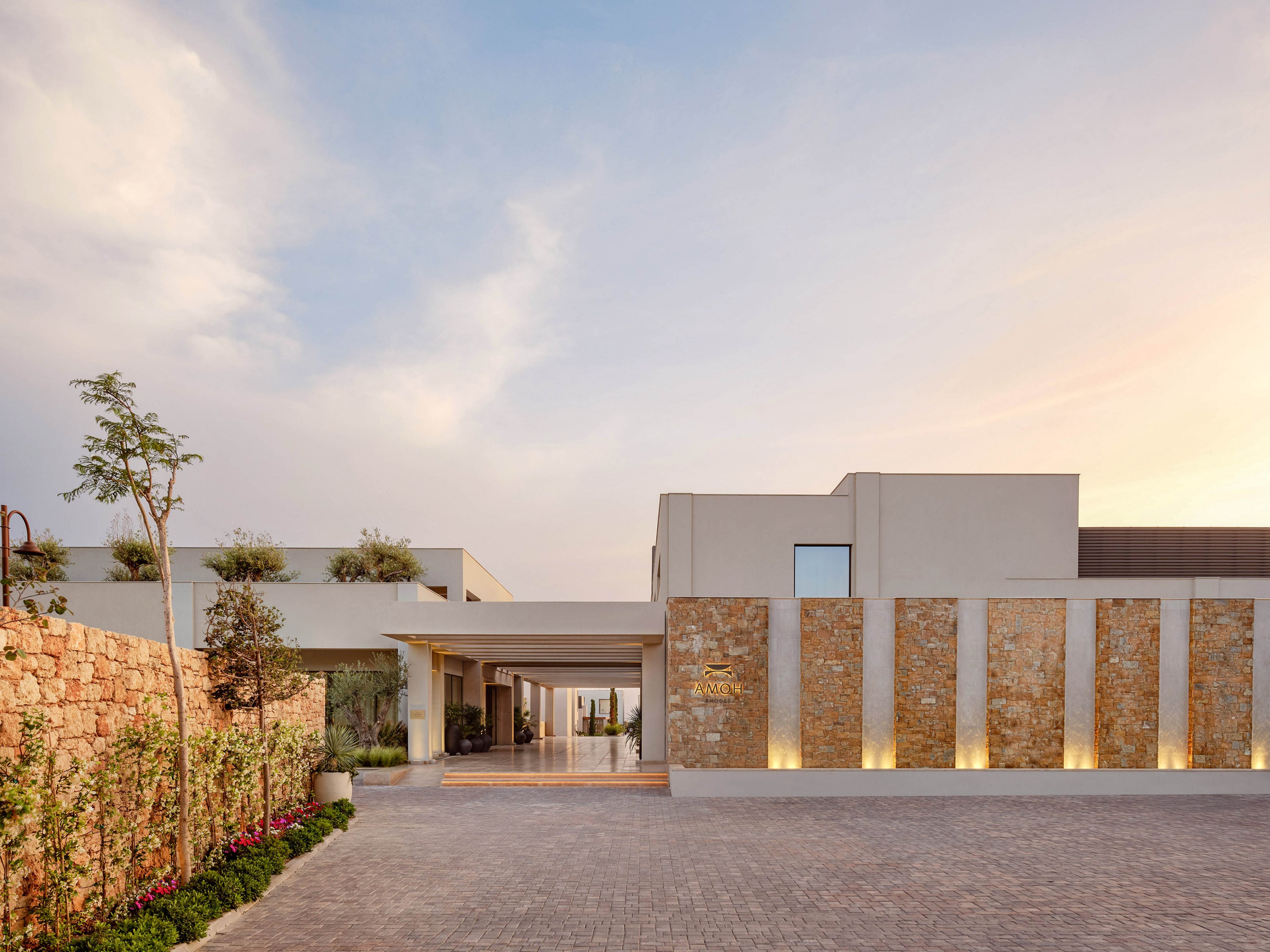 A contemporary hotel entrance features clean lines and a mix of stone and smooth plaster surfaces. Warm uplighting highlights the vertical stone columns along the facade. The wide paved driveway leads toward a covered drop-off area and glass doors. Soft evening light and a pastel sky create a calm, welcoming atmosphere, with landscaped greenery adding a touch of nature.